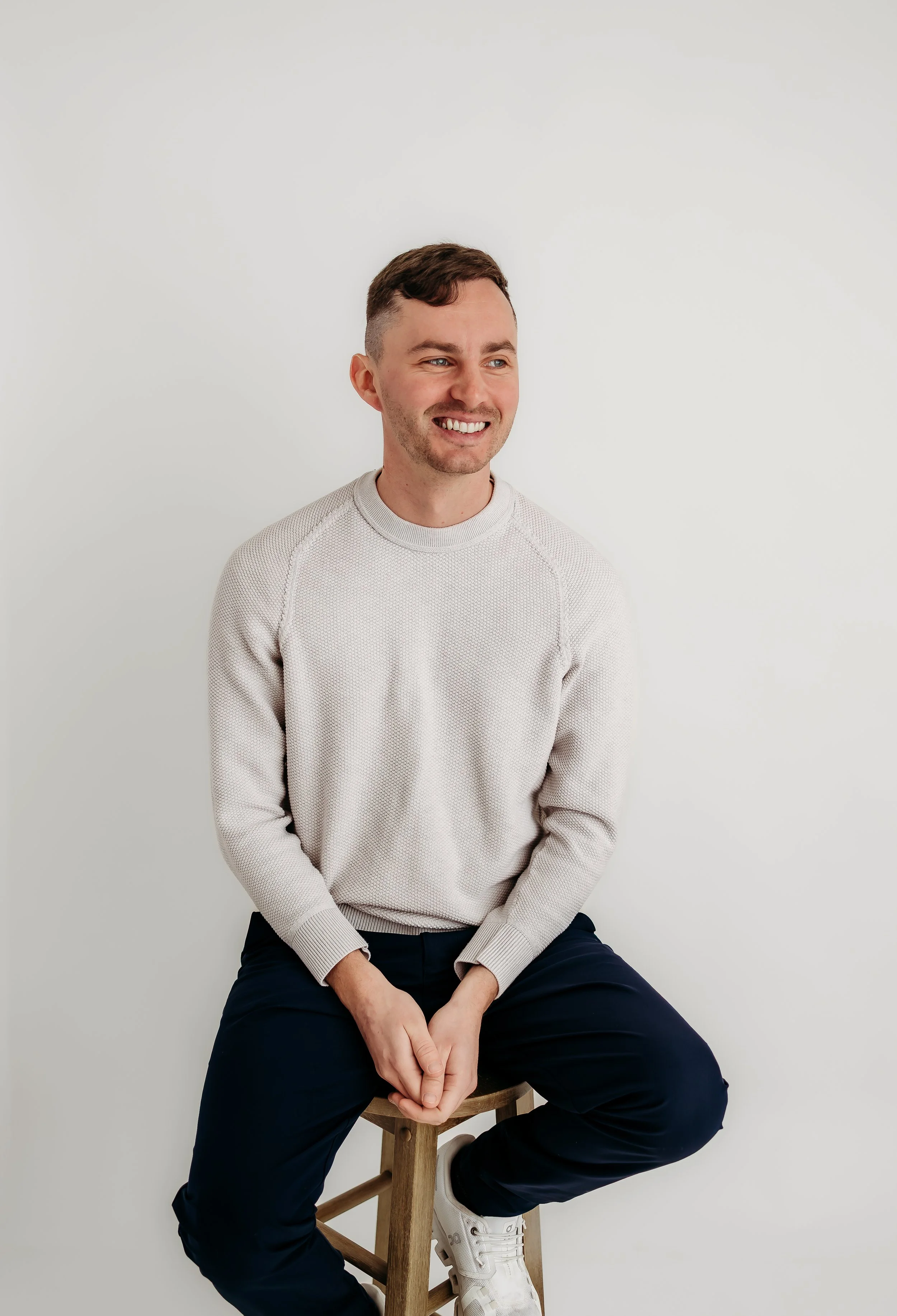 A smiling man with short brown hair, wearing a light beige sweater, dark pants, and white sneakers, sitting on a wooden stool against a plain white wall.