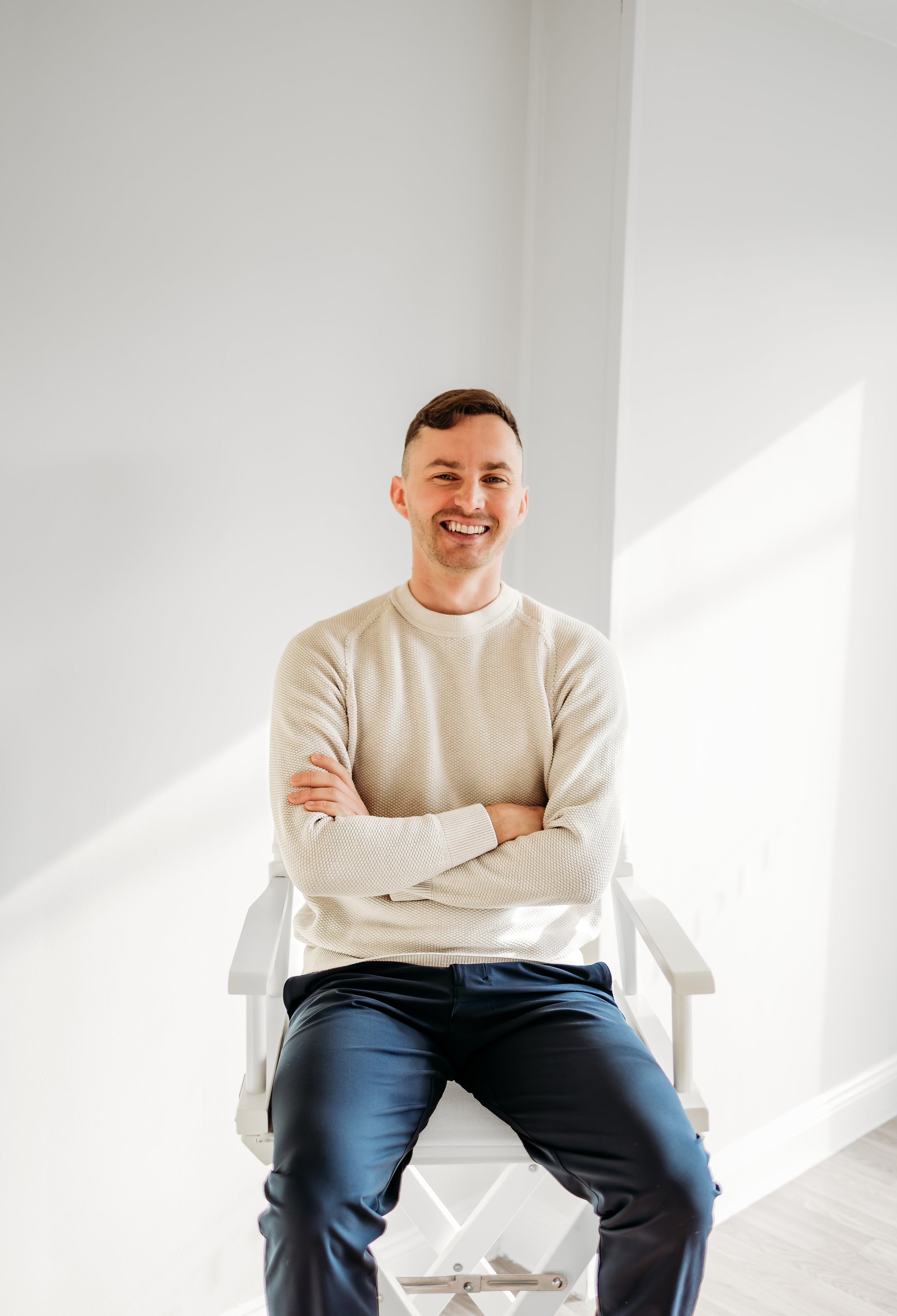 A smiling man with short dark hair and light skin, sitting on a white director's chair in a bright, minimalist room with natural light streaming in.