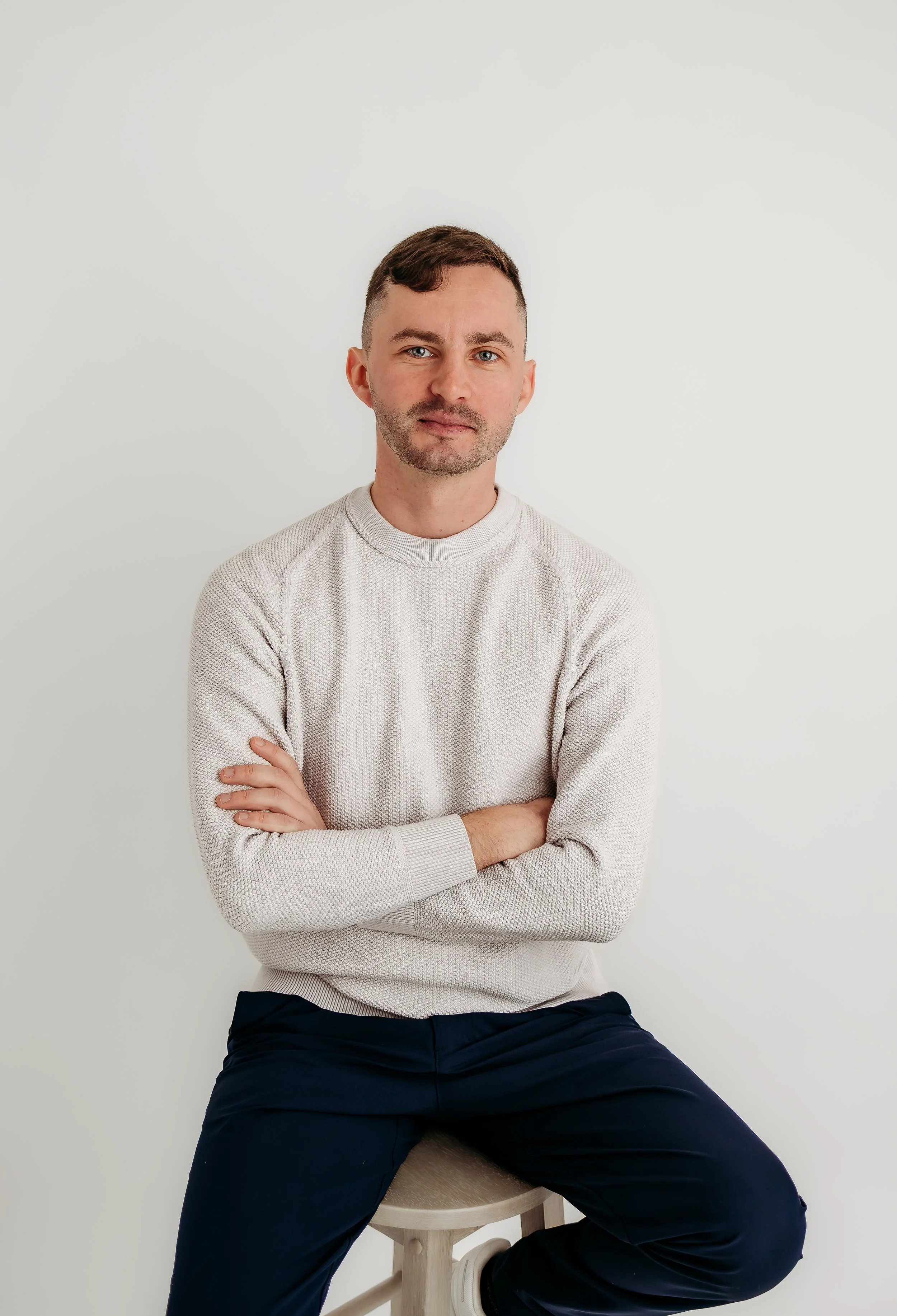 A young man with short brown hair and a beard, wearing a light gray long-sleeve shirt and dark pants, sitting on a stool with crossed arms in front of a plain white wall.