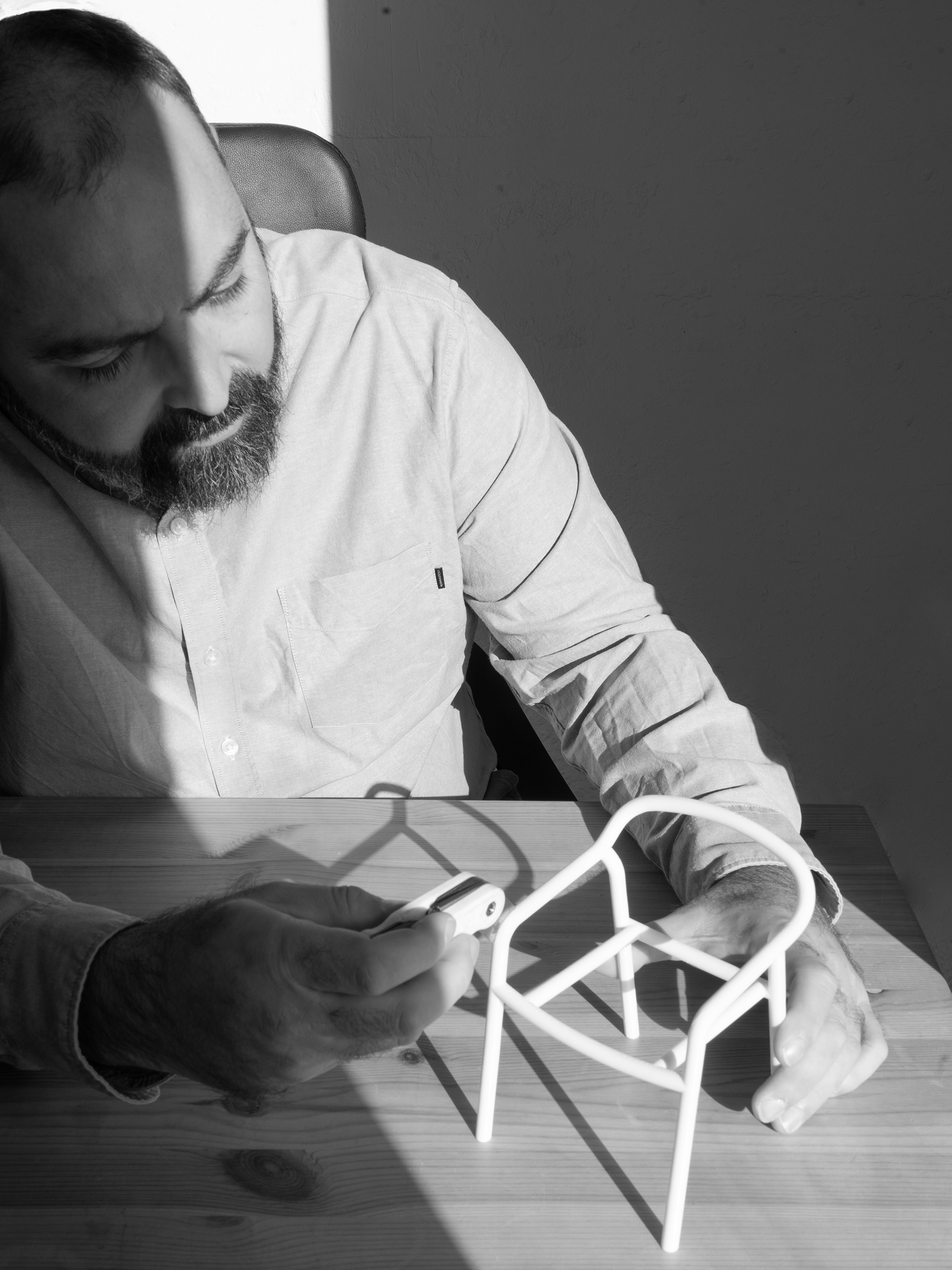 Hombre con barba vestido con camisa blanca, sentado en una mesa de madera, sostiene un dispositivo en la mano y observa una pequeña estructura de metal en la mesa.