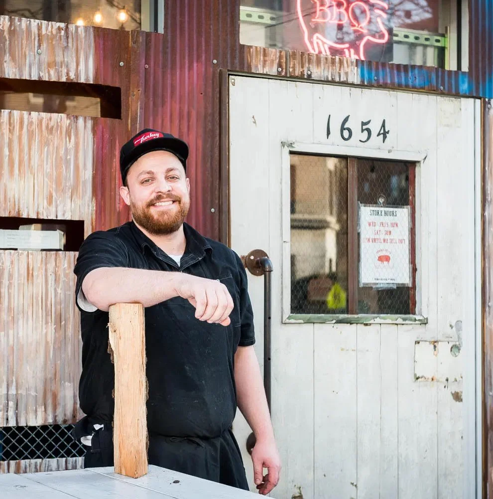 A man with a beard and a black shirt leaning on a wooden post, standing outside a rustic restaurant or shop with the number 1654 above a small window. There is a neon sign in the shape of a pig and the word 'BBQ' glowing above. The man is smiling and wearing a black cap with red and white writing.