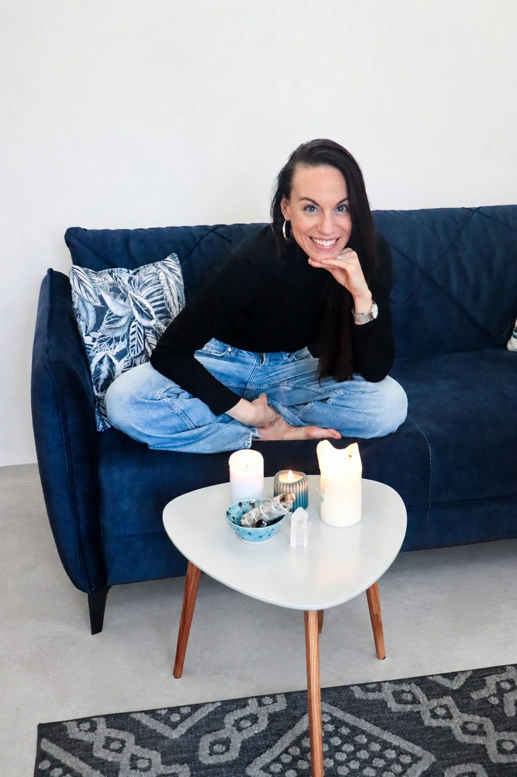 Junge Frau sitzt lachend auf einem dunklen Sofa, im Vordergrund ein kleiner Tisch mit Kerzen und Dekoration. Die Frau trägt schwarze Oberteil und Jeans, hat lange dunkle Haare, trägt Ohrstecker und Armbanduhr.