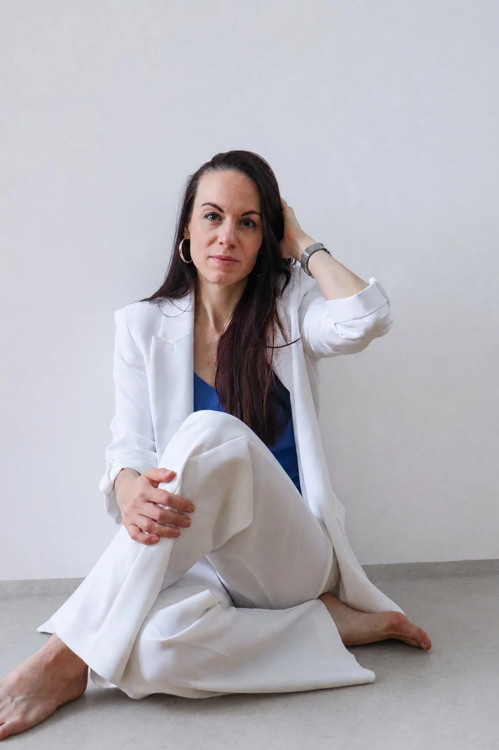 Eine Frau sitzt auf dem Boden, trägt weiße Anzugshose, eine blaue Bluse und einen weißen Blazer, mit langen braunen Haaren, die sie mit einer Hand berührt, vor einer weißen Wand.