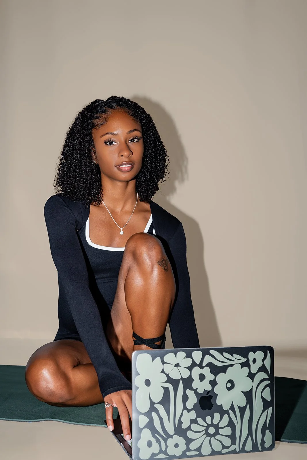 A young woman with curly black hair sitting on a Pilates mat in front of a silver laptop with floral patterns, wearing a black dress with white trim, and a necklace, in a neutral setting.