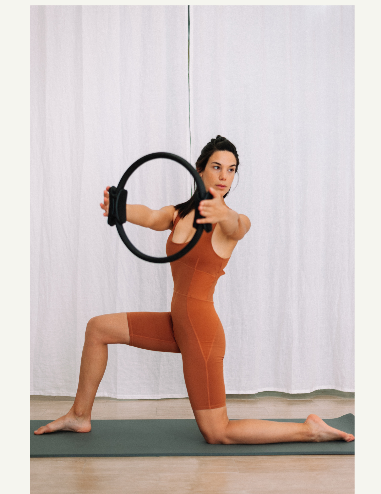 A woman doing Pilates indoors, kneeling on a mat, holding a circular yoga prop with one hand and extending her other arm forward.