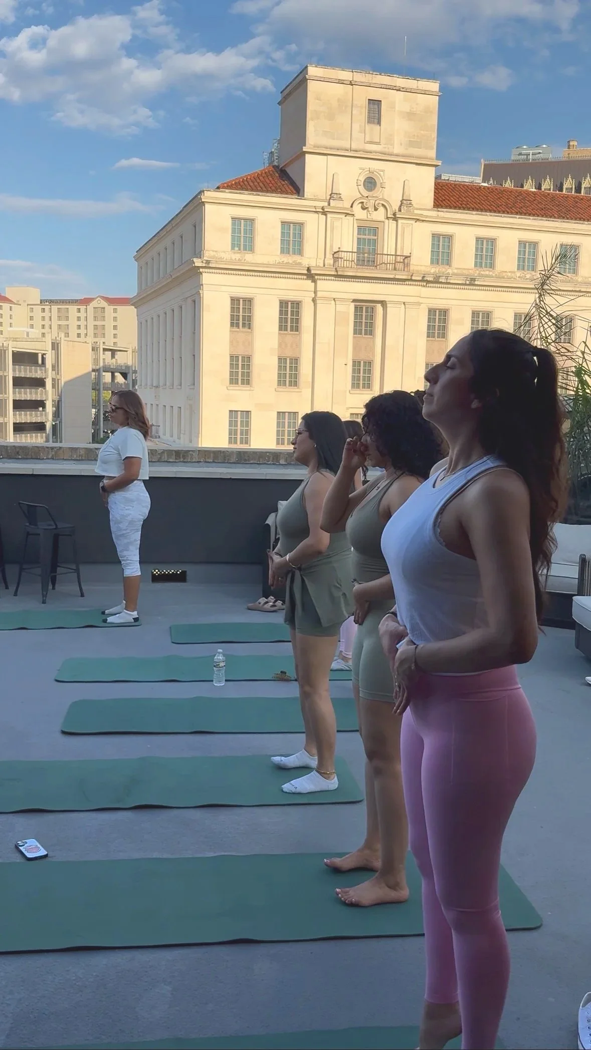Women participating in a rooftop outdoor Pilates class during the daytime with a cityscape background, some women are practicing Pilates on mats while a woman in white appears to be instructing.