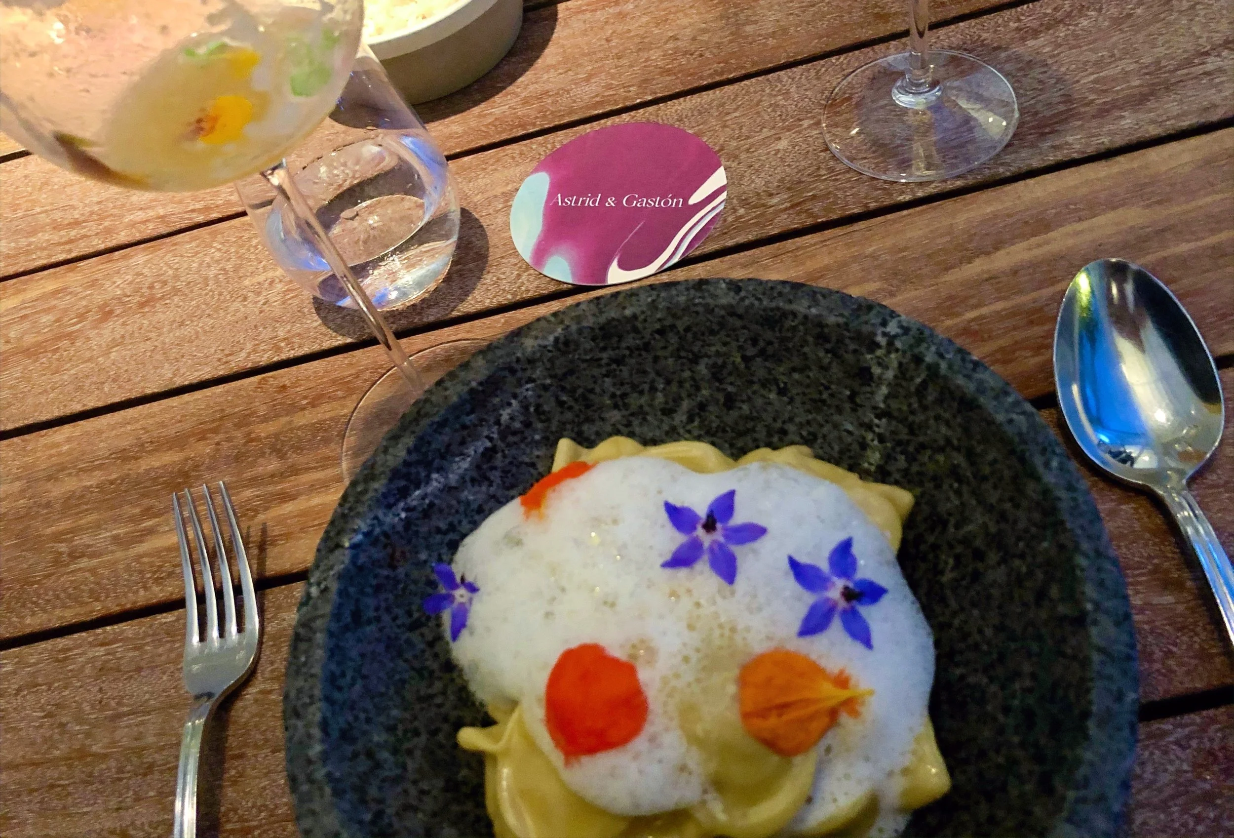 A plate of pasta with edible flowers on top, set on a wooden table with 'Astrid & Gastón' on a coaster in the background.