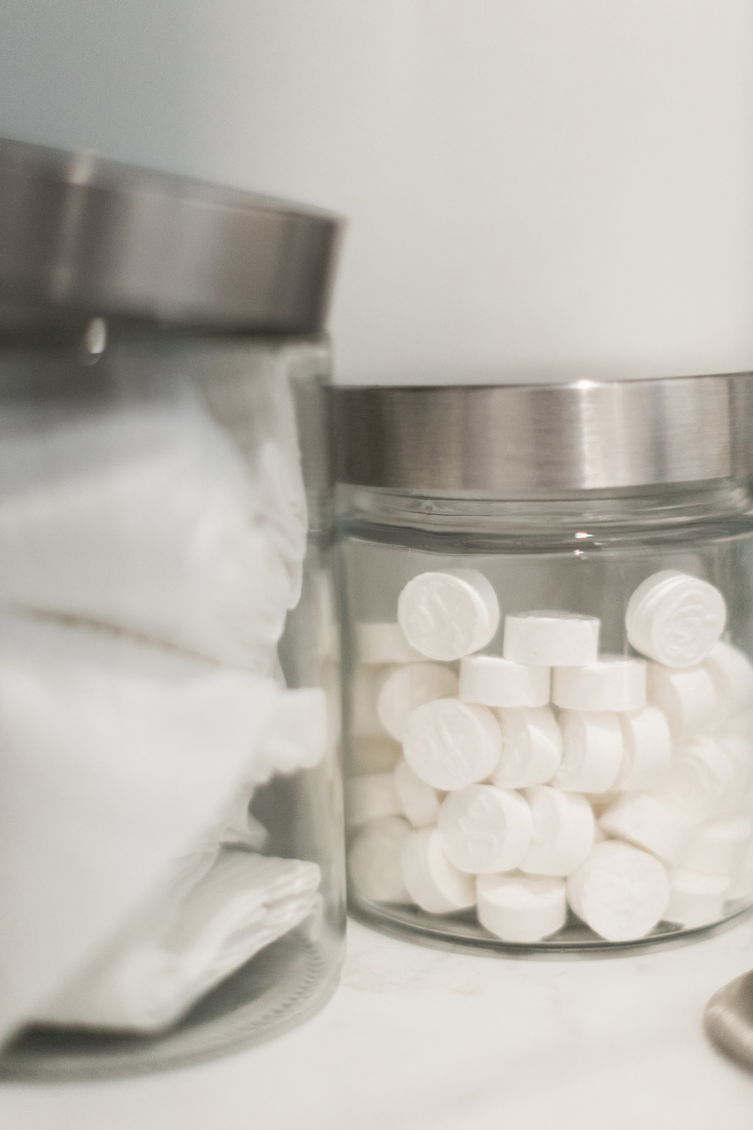 Close-up of a glass jar filled with white, round pills or tablets next to a larger jar containing white powder, both on a white surface.