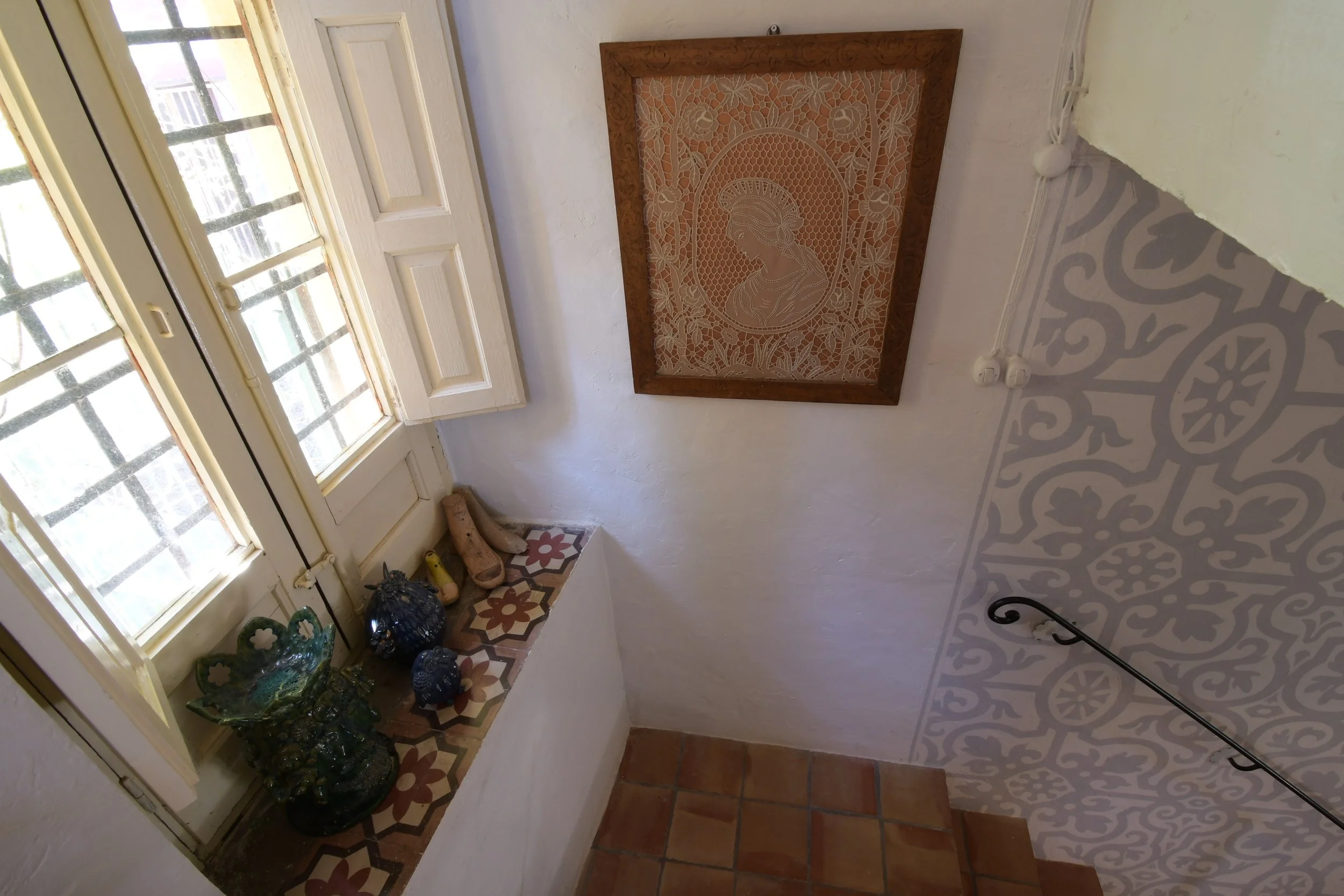 View of an entryway with tile flooring, a decorative framed embroidery of a woman's profile, a small window with bars, and a collection of ceramic and metal decorative items near the window.