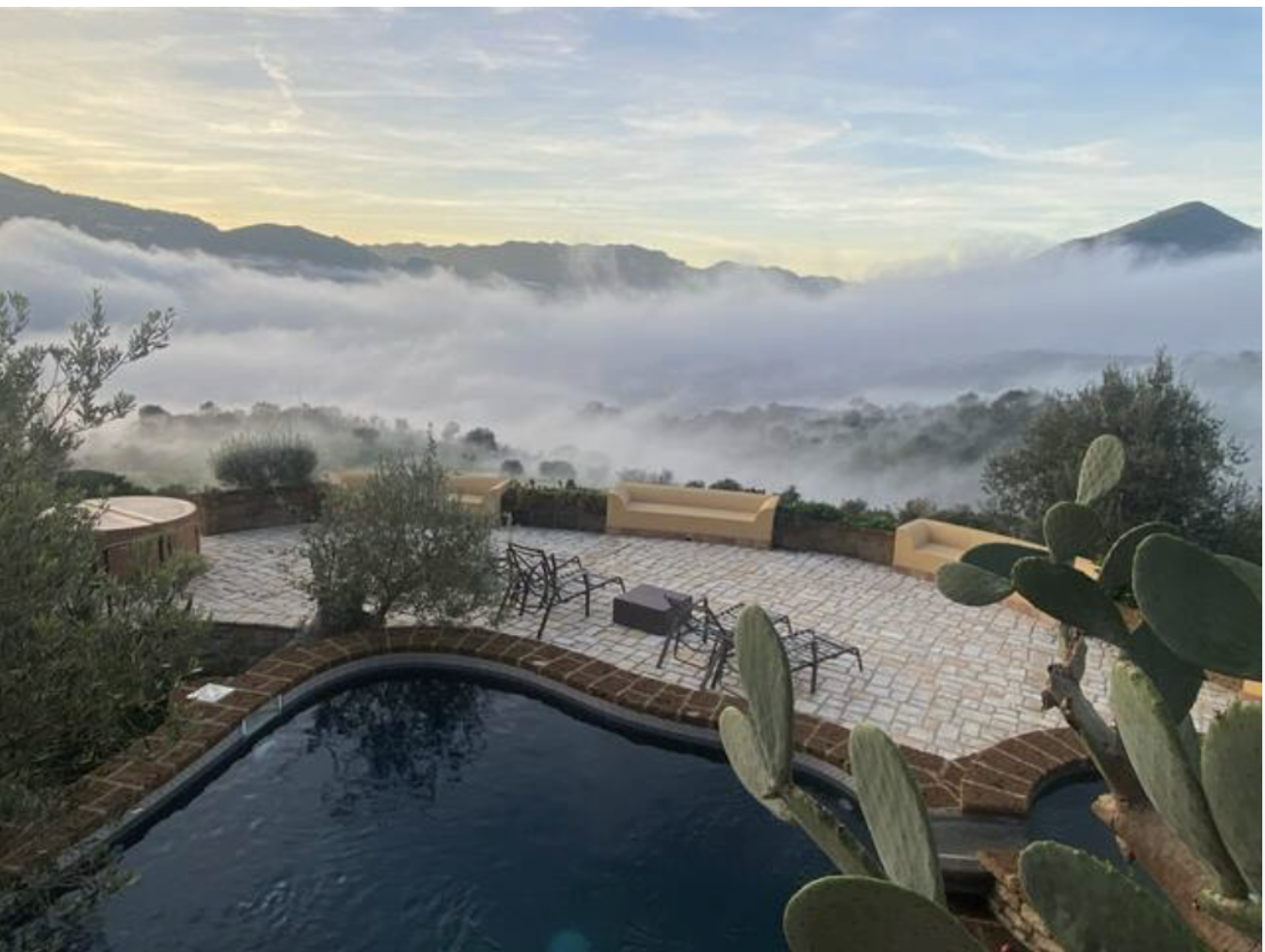 A scenic outdoor patio with a hot tub, lounge chairs, and benches overlooking a foggy valley with mountains in the background, under a partly cloudy sky.