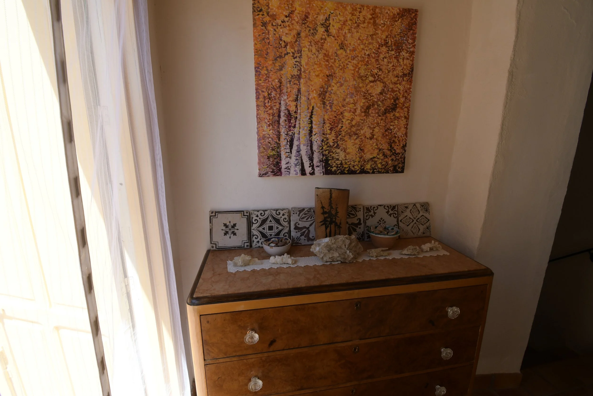 A wooden dresser with a marble top in a room with a painting of an autumn landscape on the wall, decorative bowls and stones on the dresser, and a window with curtains on the left side.