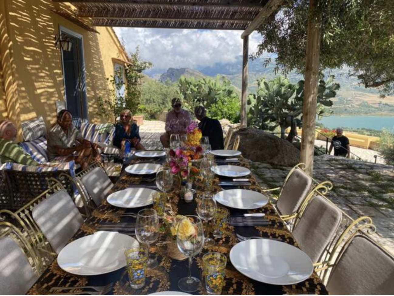 Outdoor dining table set with plates, glasses, and a floral centerpiece, with people seated on a patio overlooking a hillside and water, shaded by a pergola.