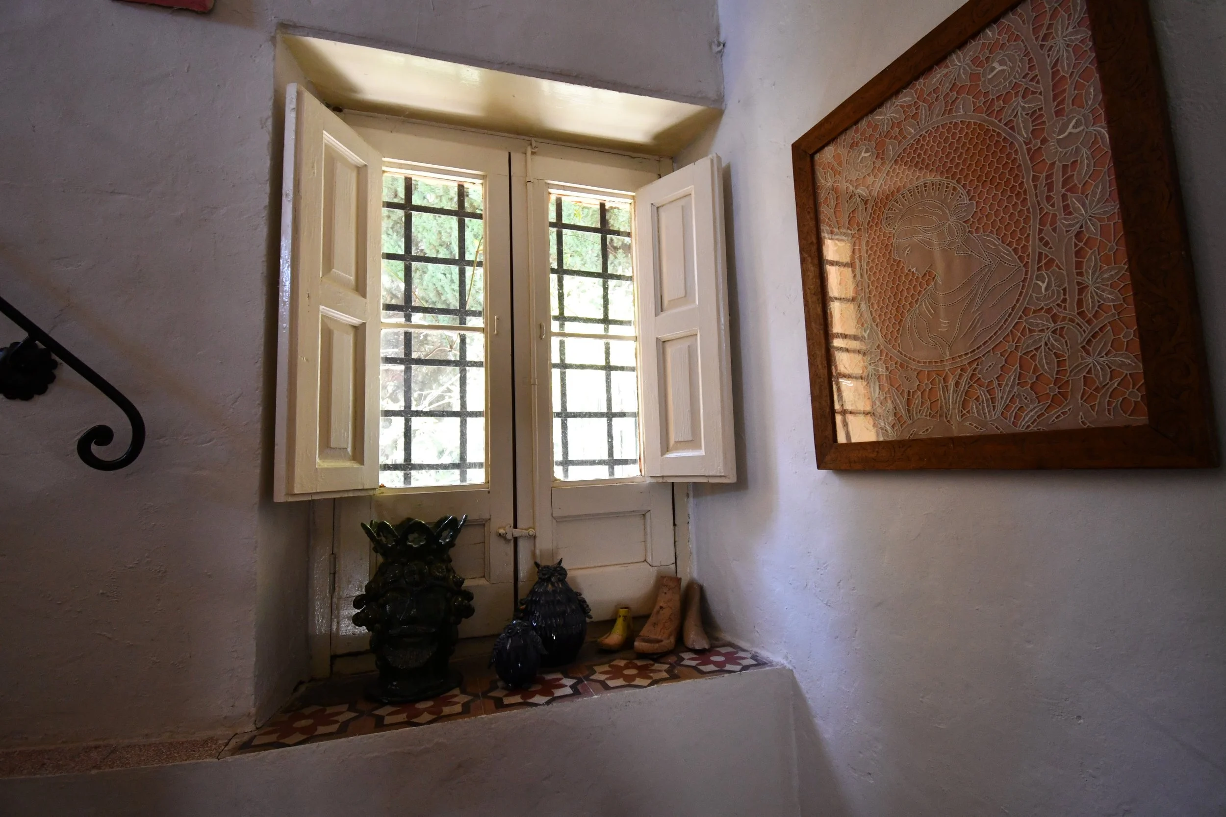 A window with open cream-colored shutters and a metal grille, set in a white stucco wall, with decorative tile flooring and a large framed artwork with an intricate design leaning against the wall nearby.