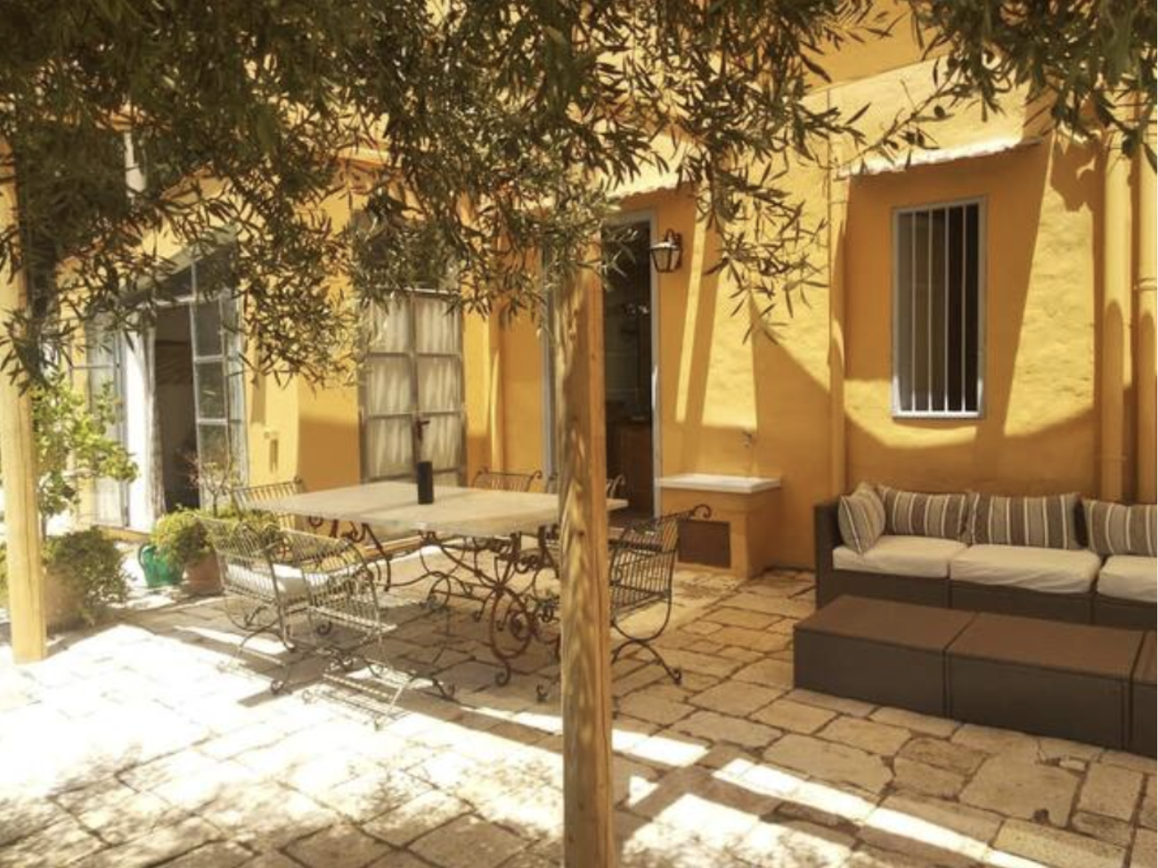 A sunlit outdoor patio with a beige stone tile floor, enclosed by a yellow stucco wall. There is a large rectangular dining table with a bottle on top, surrounded by ornate wrought iron chairs. To the right, a cushioned outdoor sofa with striped pill