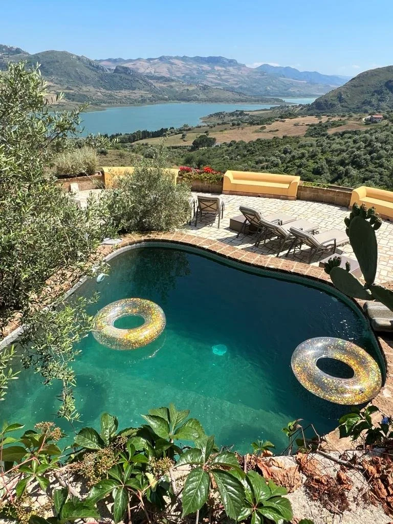 A swimming pool with two inflatable rings floating on the water, overlooking a scenic view of a lake, mountains, and a landscaped patio with lounge chairs and benches surrounded by greenery.