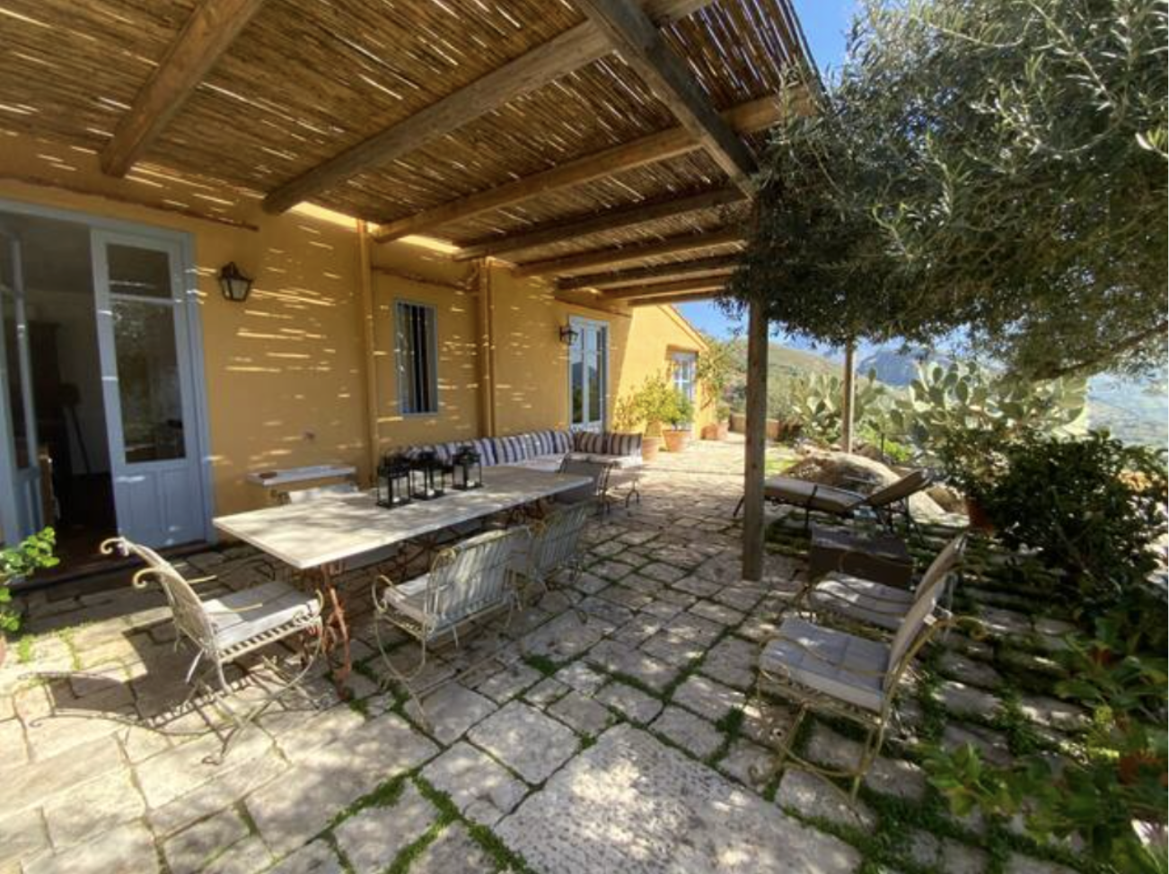 Outdoor patio with stone flooring, metal chairs, a dining table with lanterns, a yellow wall with windows, potted plants, and a pergola with wooden slats casting shadows. There's a view of greenery and hills in the background.