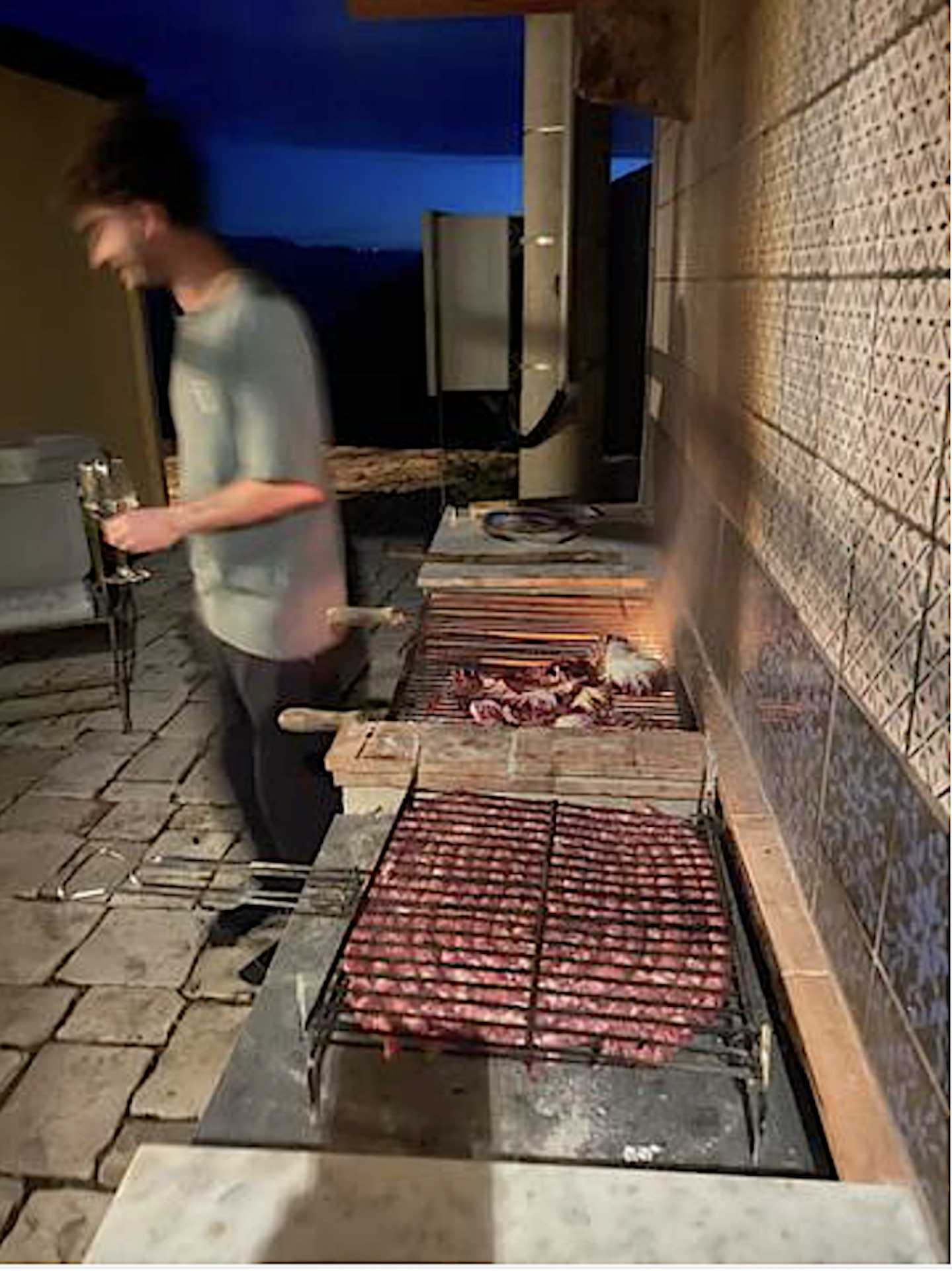 A person grilling meat on an outdoor barbecue grill at dusk or night. The scene includes a brick patio, a table with a glass of water, and a wall with patterned tiles.