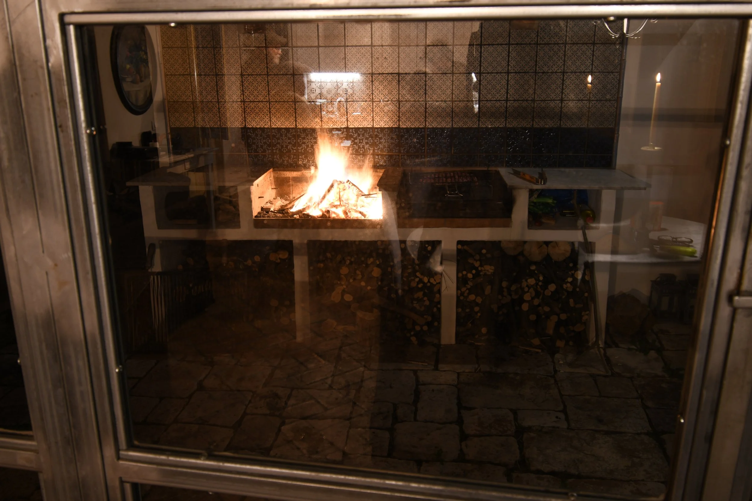 A view through a glass window of an indoor fireplace with a fire burning, surrounded by tiled walls and a countertop with kitchen tools and a stack of logs underneath.