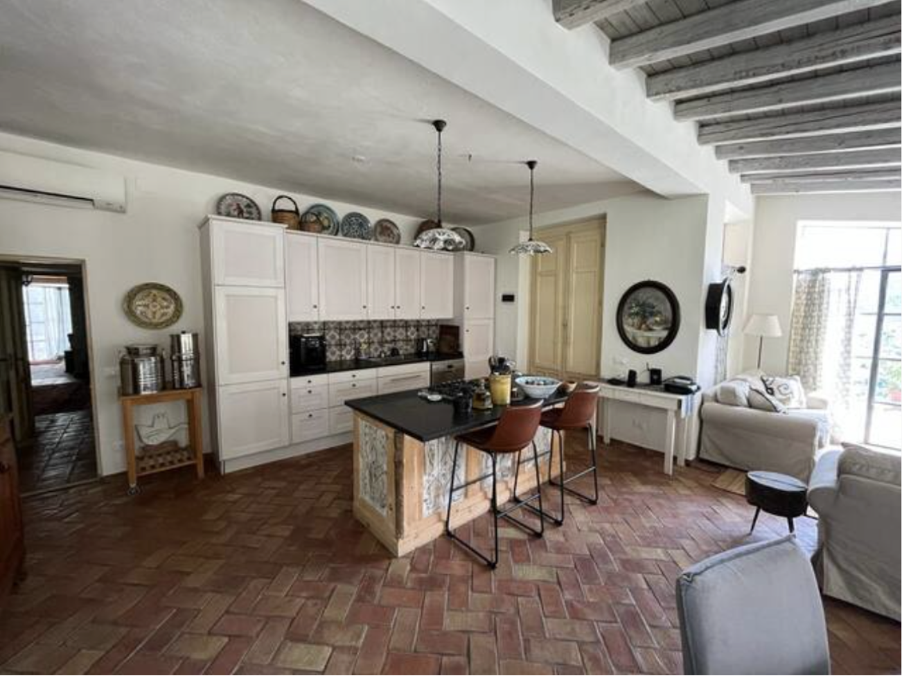 Open-concept living room and kitchen with white cabinets, a small kitchen island, terracotta tile flooring, white sofas, and large windows.