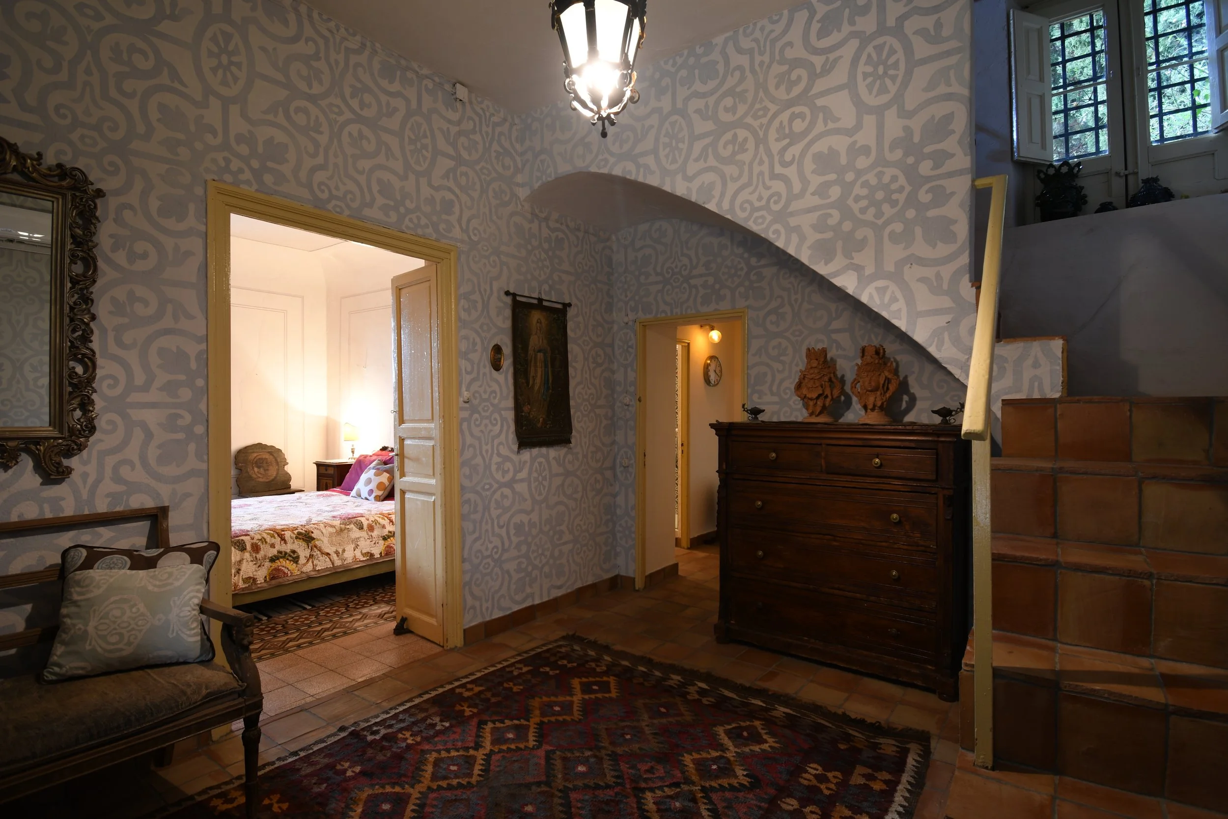 Living room with a staircase, vintage furniture, and a bedroom visible through an open doorway. Decor includes a patterned rug, ornate mirror, and various wall hangings, with natural light coming from a window.