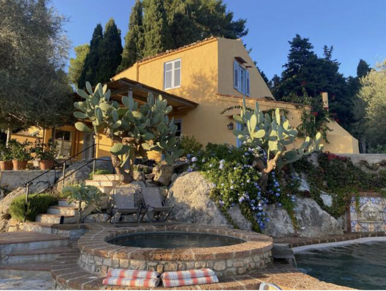 A yellow house with white windows is situated on a hillside surrounded by tall cypress and other trees. In the foreground, there is a rock garden with large prickly pear cacti, flowering plants, a hot tub, and a swimming pool. Sunlight and shadows su