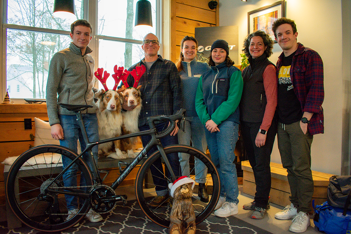 Group of seven people standing inside near a window, with two dogs wearing reindeer antlers and a bicycle with a Santa hat on a carved dog statue in the foreground, during a holiday celebration.