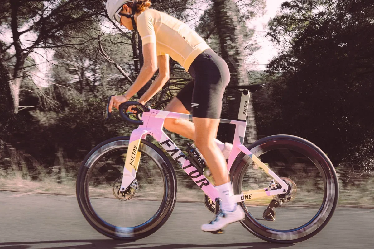 A woman riding a pink Factor racing bicycle on a paved road in a wooded area, wearing a helmet, yellow cycling jersey, black shorts, and white shoes.