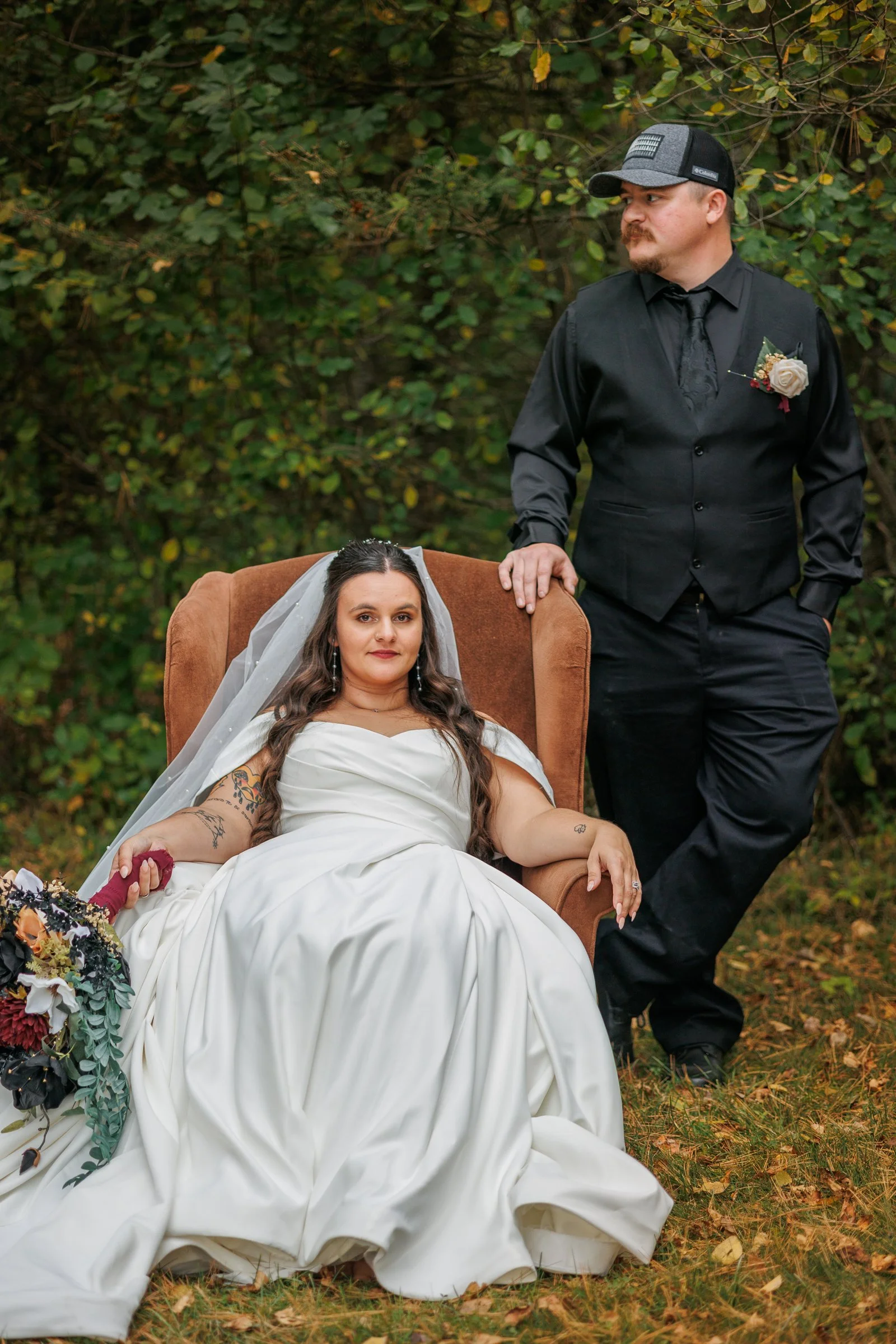 Bride in white dress sitting on a chair outdoors, holding a bouquet, next to a man in dark attire and hat standing, with a wooded background.