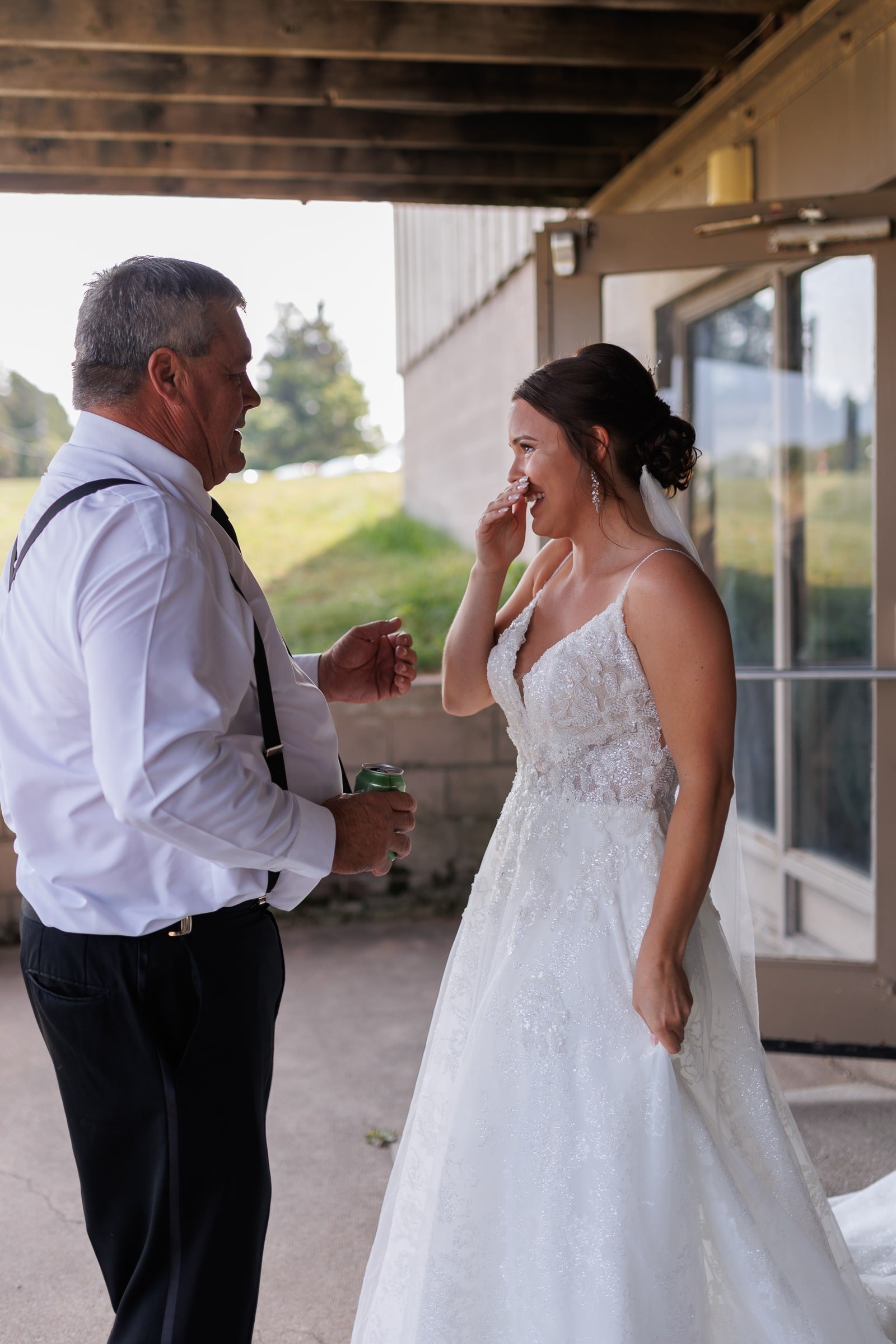 Bride in a white gown smiling and touching her face, standing with a man in a white shirt and suspenders, outdoors with a green lawn and glass door in the background.