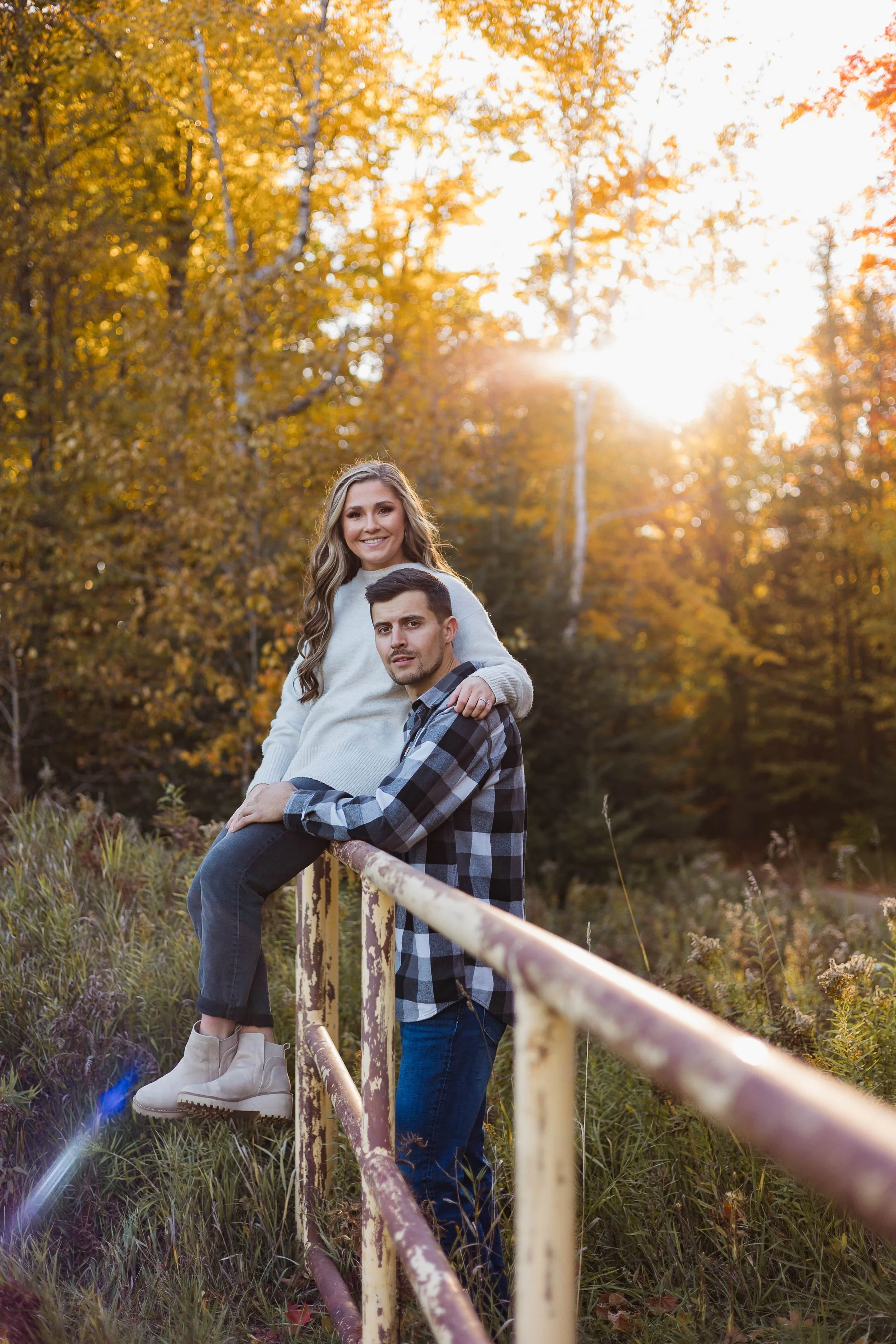 Couple posing on rustic railing in autumn forest with sunlight through trees.