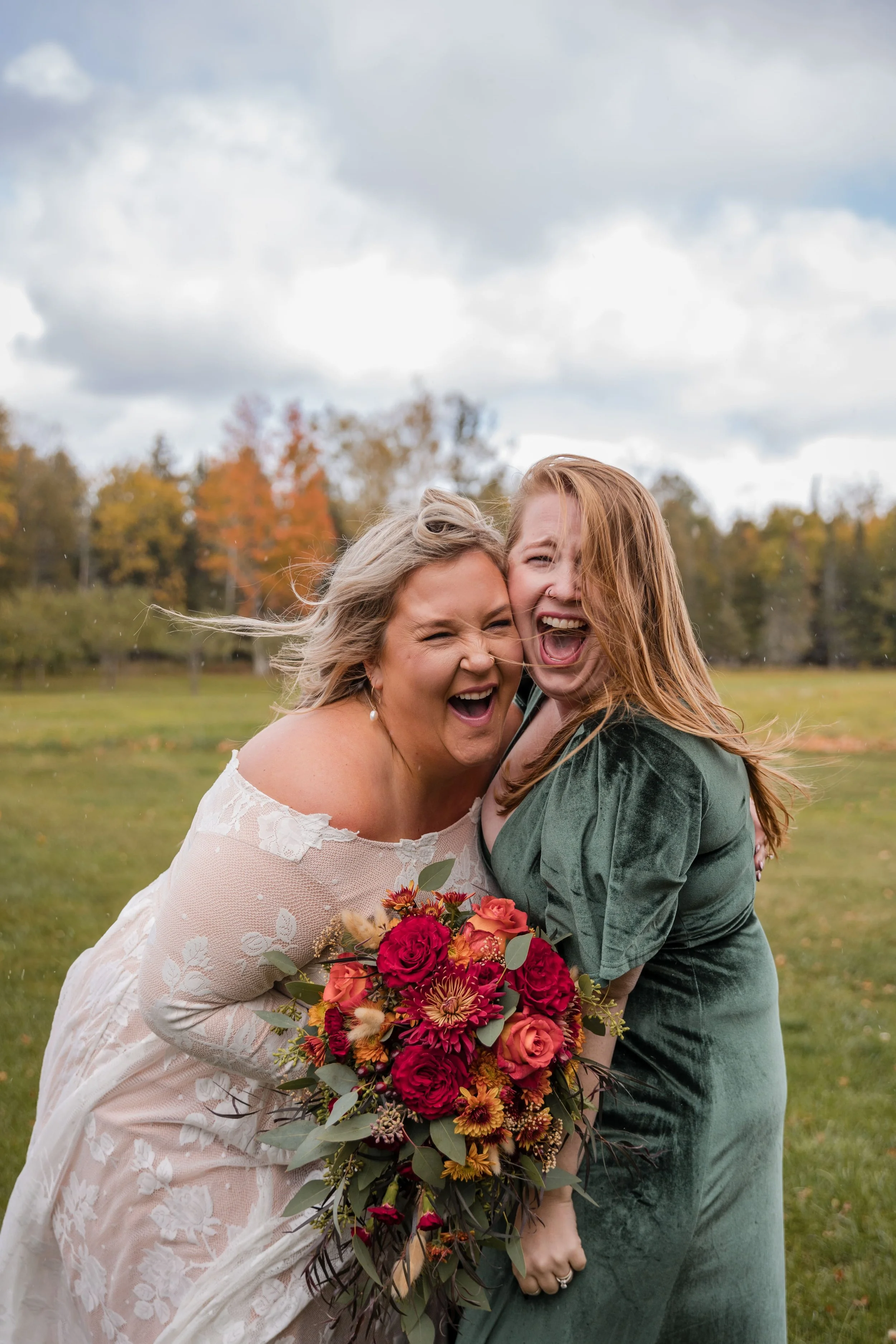 Two women laughing, one in a white lace dress holding a bouquet, the other in a green dress, standing in a field with autumn foliage.