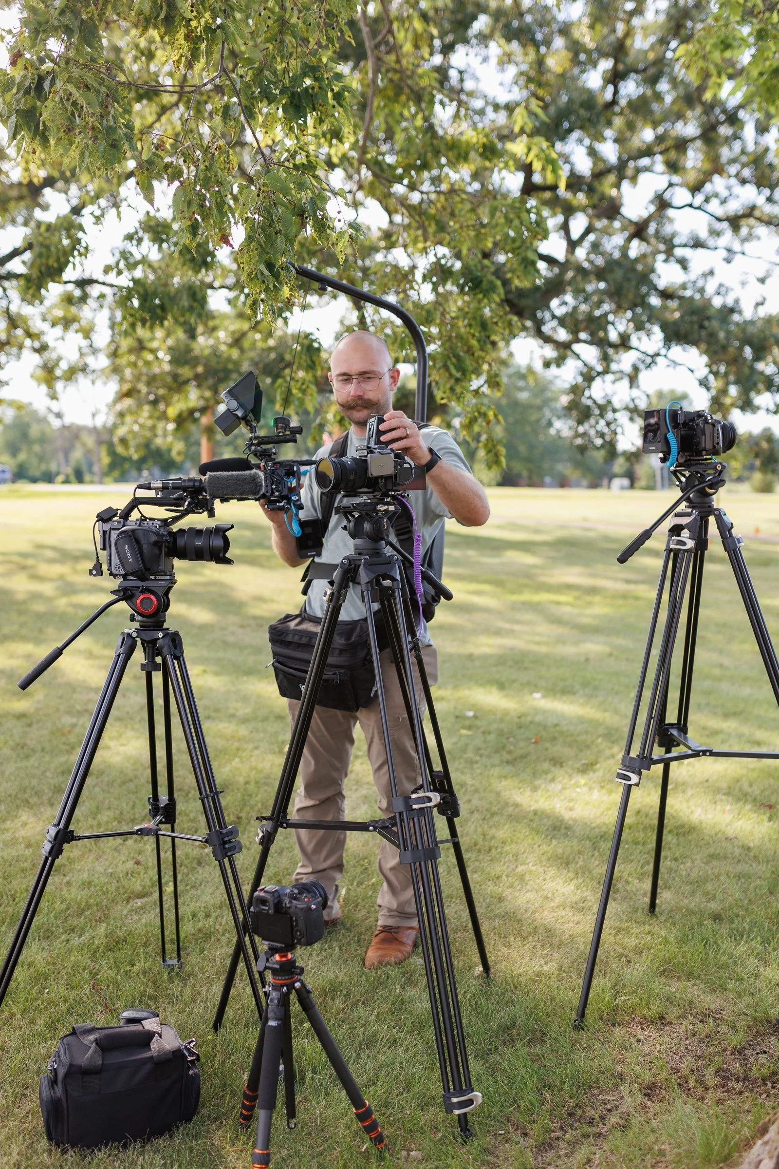 A man with glasses, a beard, and a mustache working with video cameras on tripods in a park, under a leafy tree.