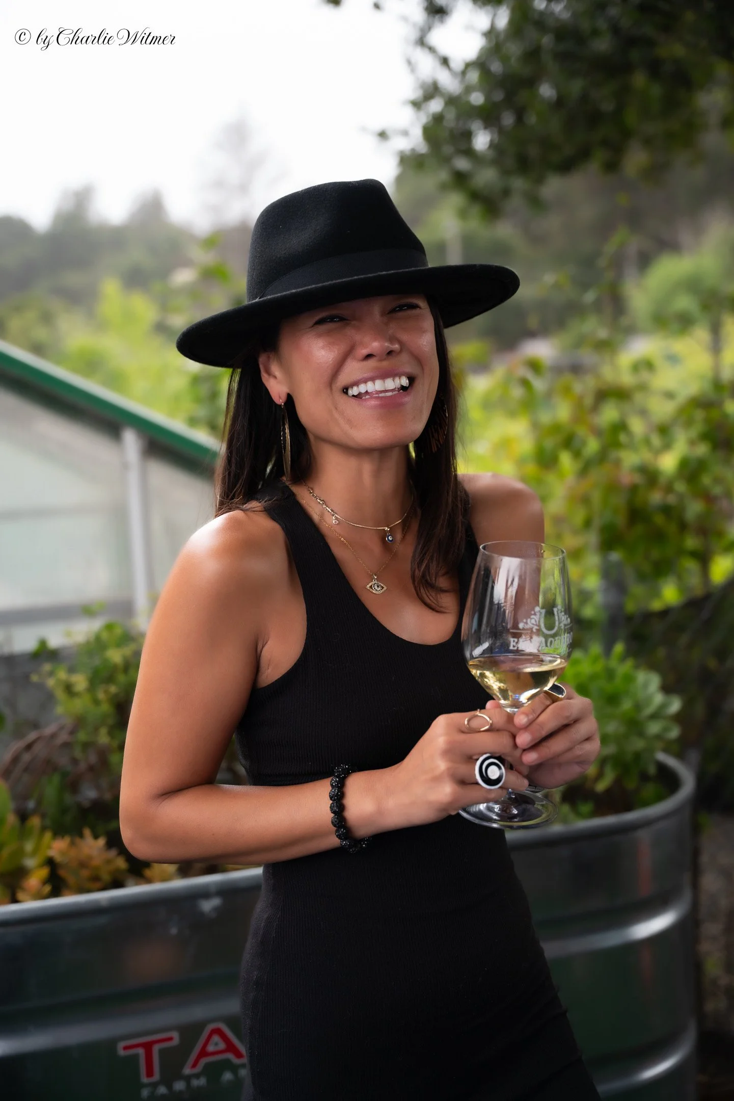 A woman in a black sleeveless dress and black hat is smiling and holding a glass of white wine outdoors, with greenery in the background.