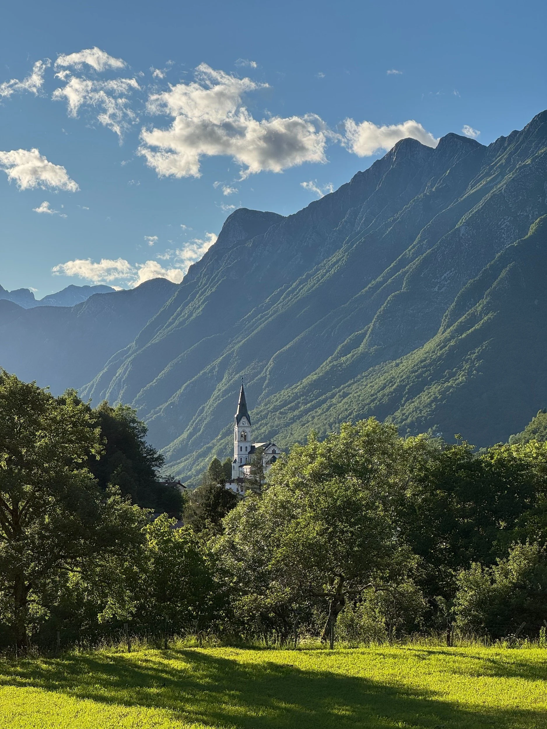 in the image you can see the church of Drežnica in Slovenia with mountains in the background, trees around the church and a lush meadow in the front