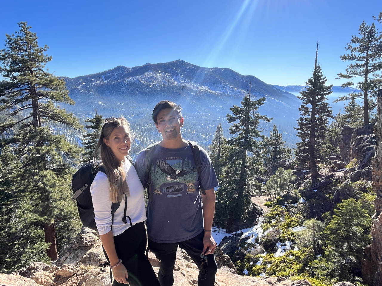 A smiling couple with backpacks hiking in a forested mountain area with snow and evergreen trees, bright blue sky, and sun rays.