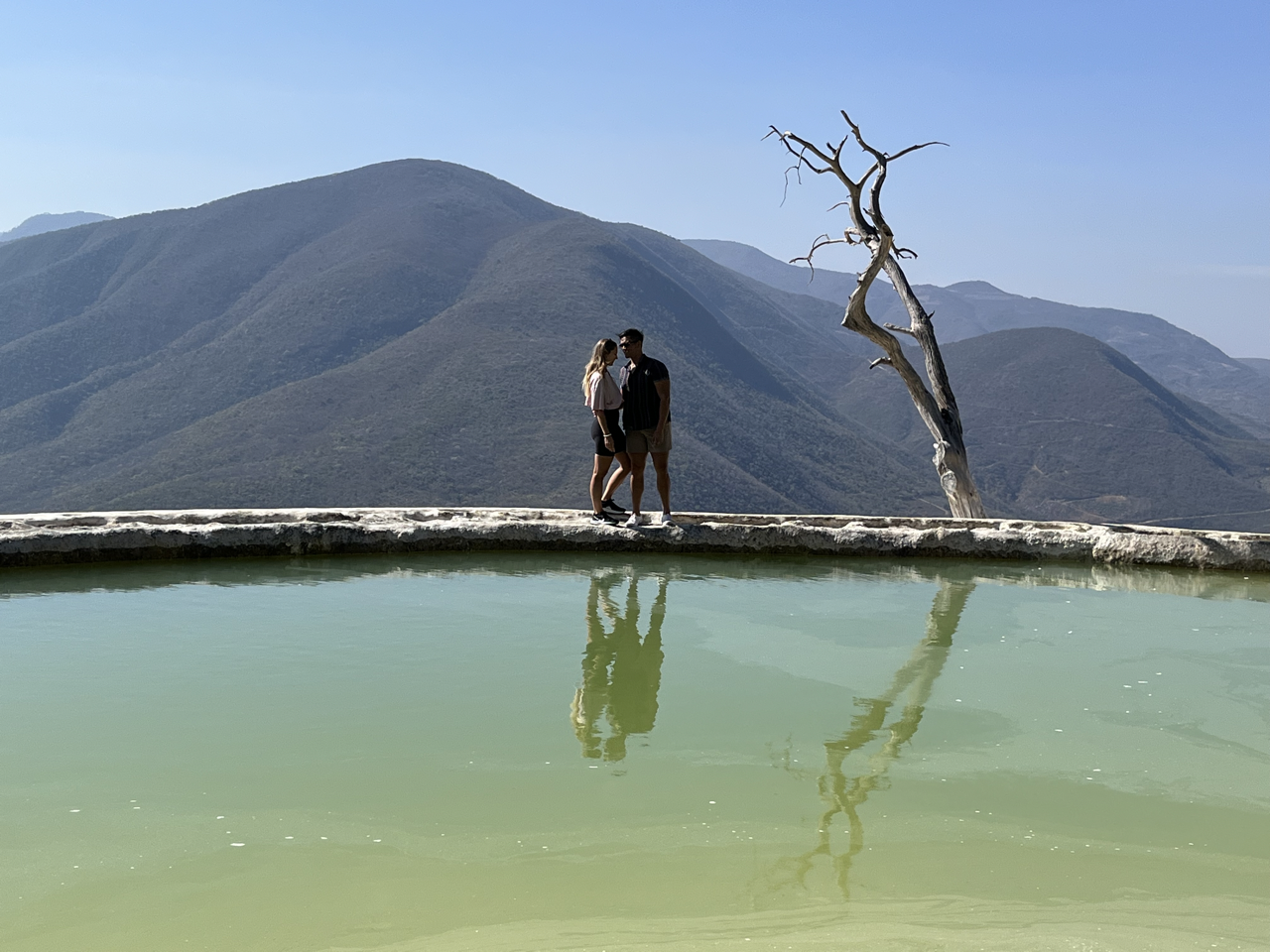 A couple standing on top of a petrified waterfall in Mexico near Oaxaca, with mountains in the background, a leafless tree, and clear blue sky.