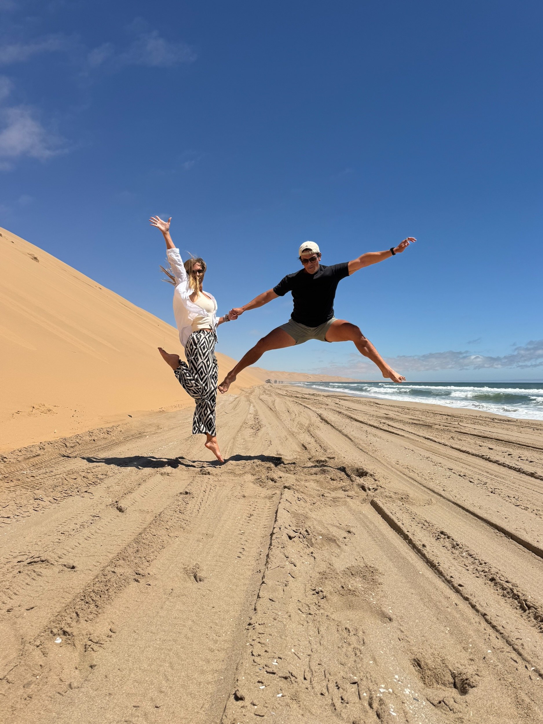 Nora and Trevor jumping beside the sand dunes and Atlantic Ocean at Sandwich Harbour, Namibia.