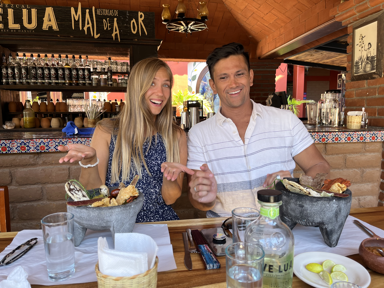 A happy young woman and man sitting at a restaurant table with Mexican food in stone bowls, glasses of water, and a bottle of mezcal. The restaurant has warm brick walls, shelves with bottles, and colorful decor.