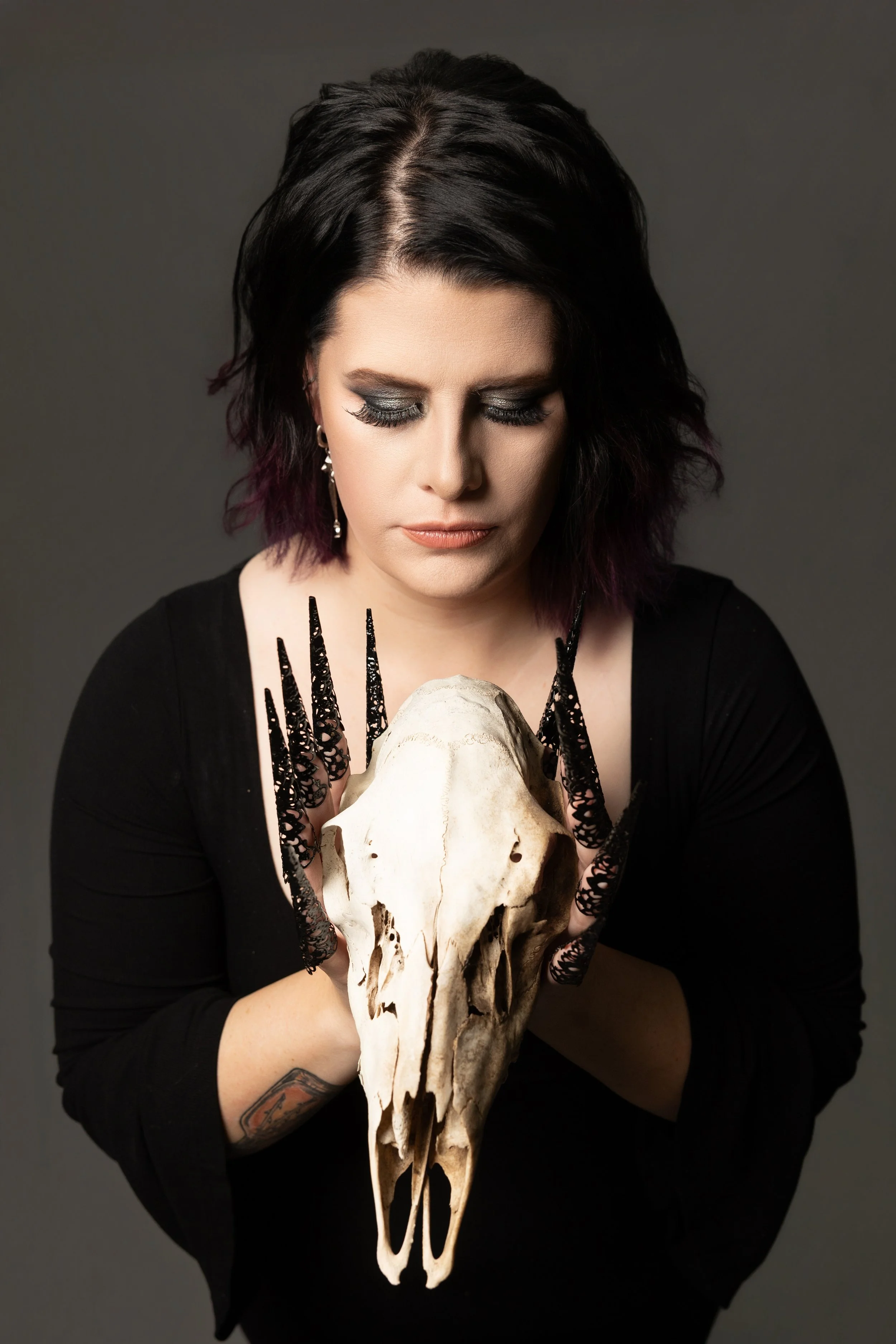 A woman with dark hair and makeup holding a large animal skull with long black claws. She is wearing a black top and earrings, with a neutral background.