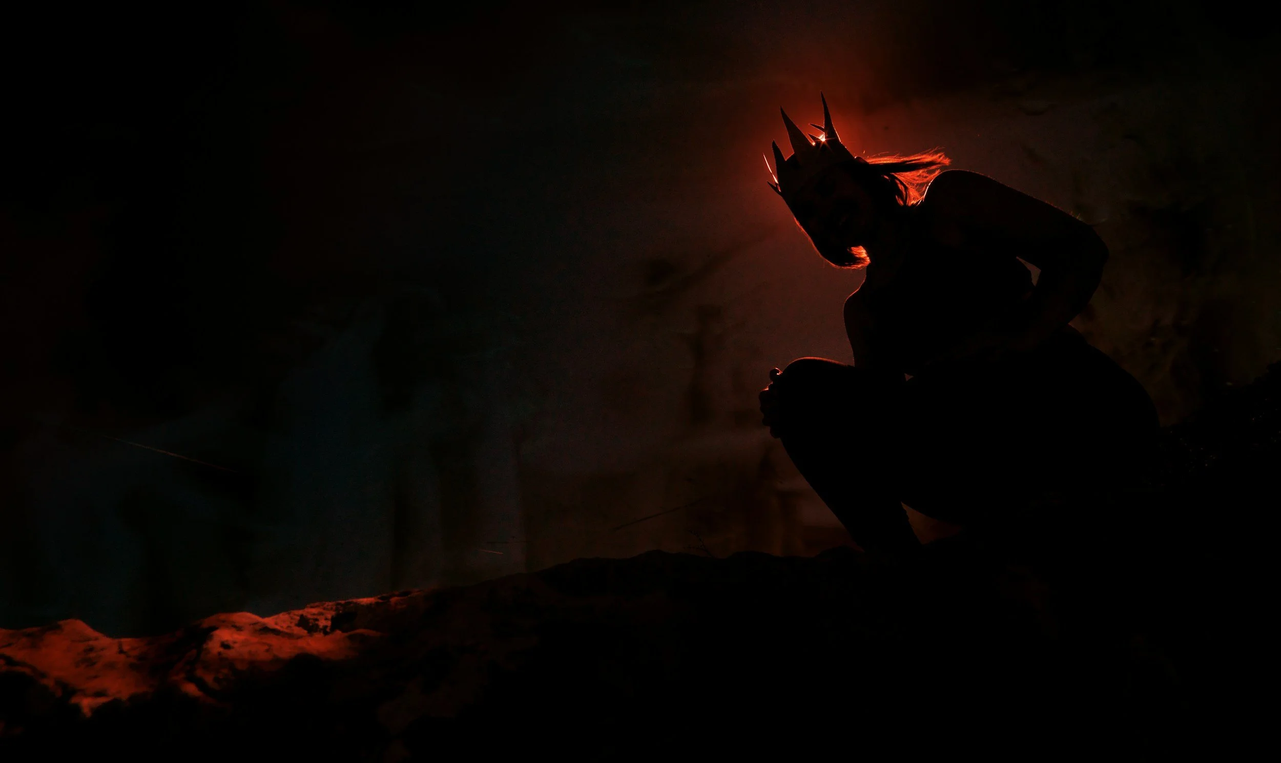 Silhouette of a woman wearing a crown, crouching on a mountain edge at sunset with a dark sky and orange glow.