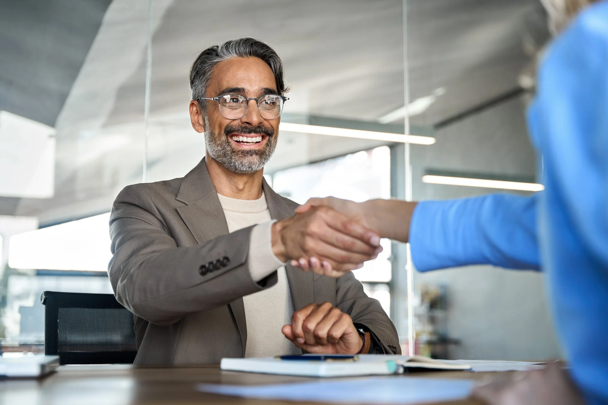 A smiling man with gray hair and beard, wearing glasses and a beige blazer, shaking hands with an unseen person in a blue blazer in an office setting.