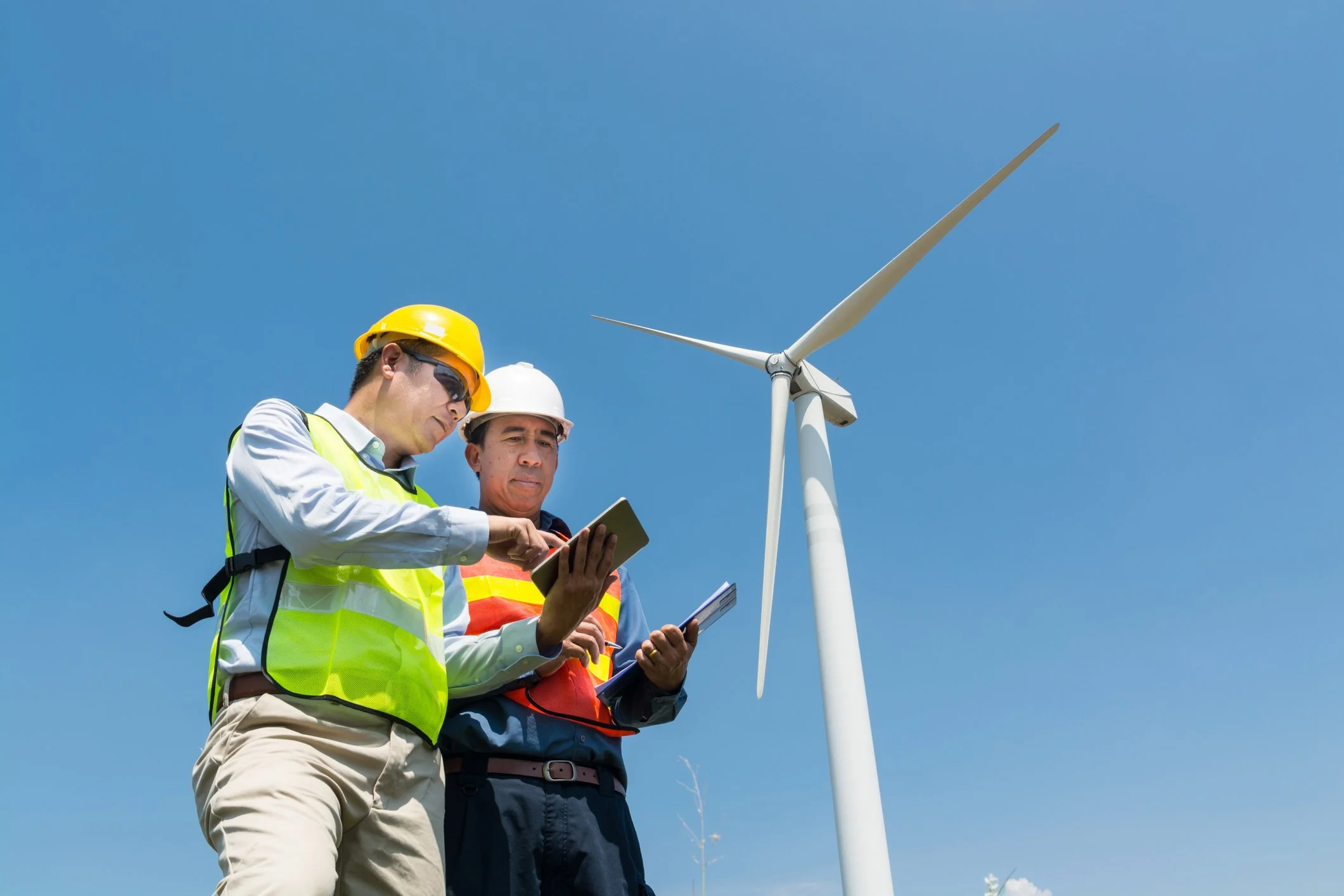 Two engineers wearing safety vests and helmets inspecting a wind turbine under a clear blue sky.