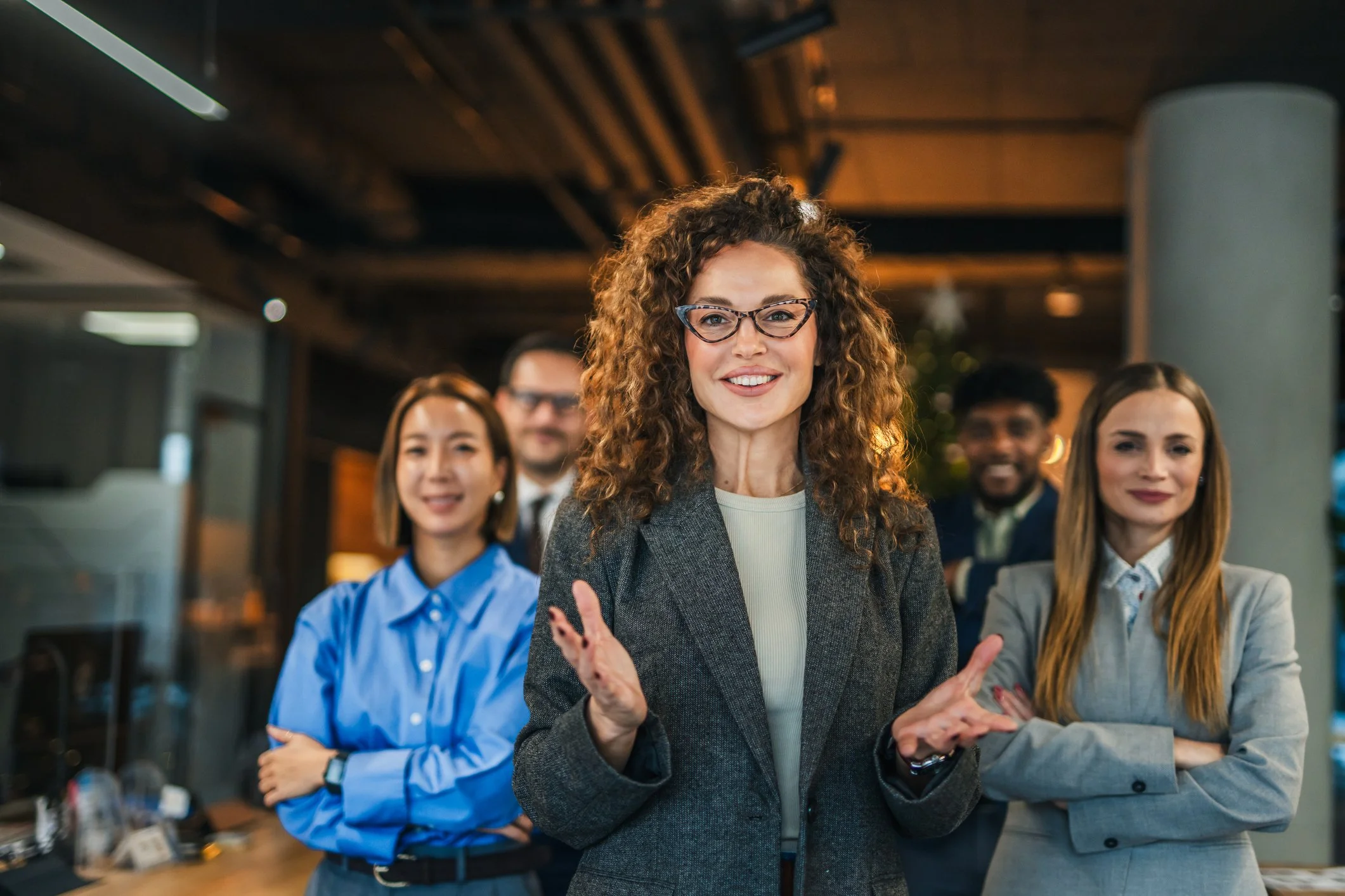 Group of diverse professionals standing in an office, smiling, with the central woman wearing glasses and gesturing with her hands.