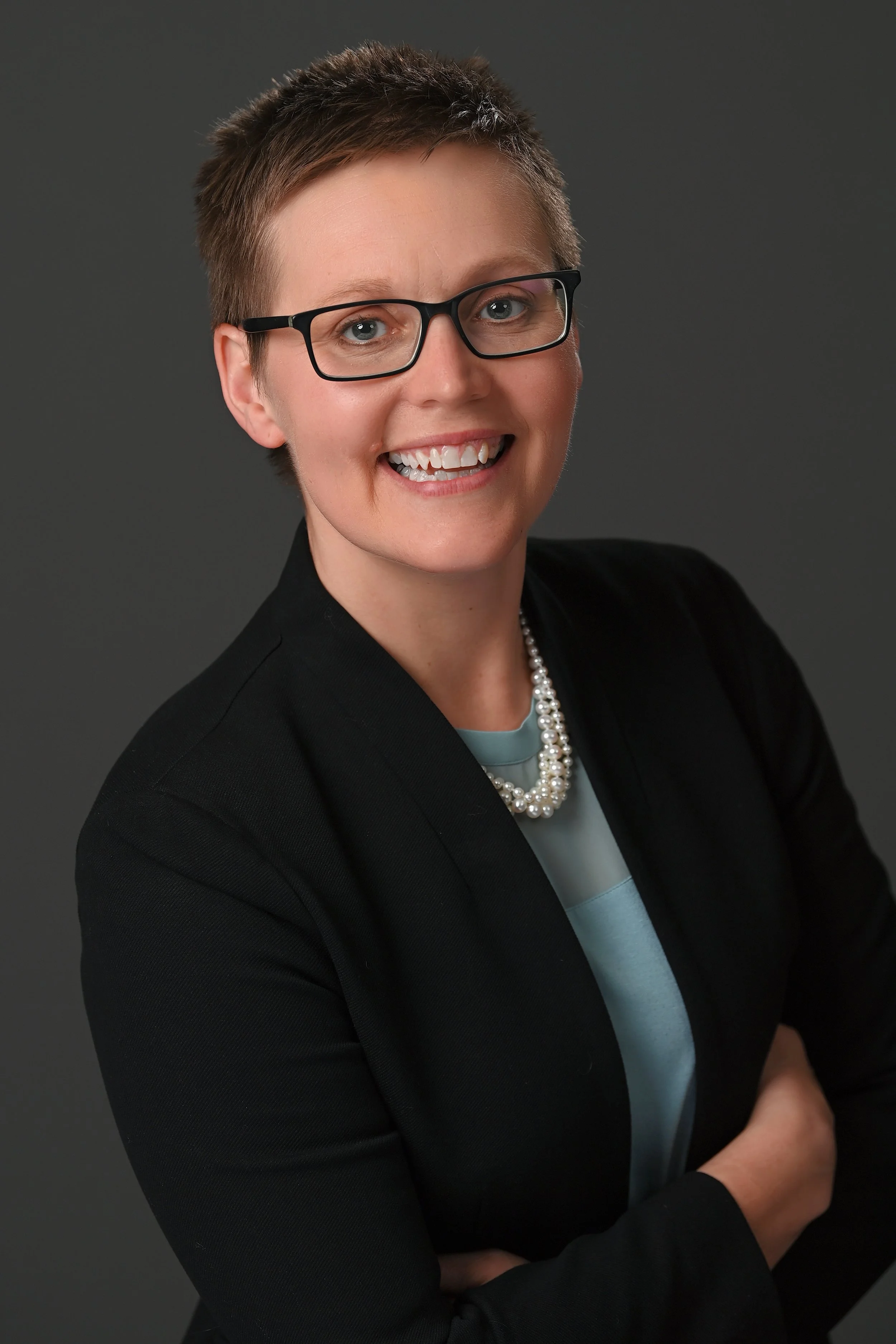 A professional woman with short hair, glasses, wearing a black blazer, light blue blouse, and a pearl necklace, smiling against a dark background.