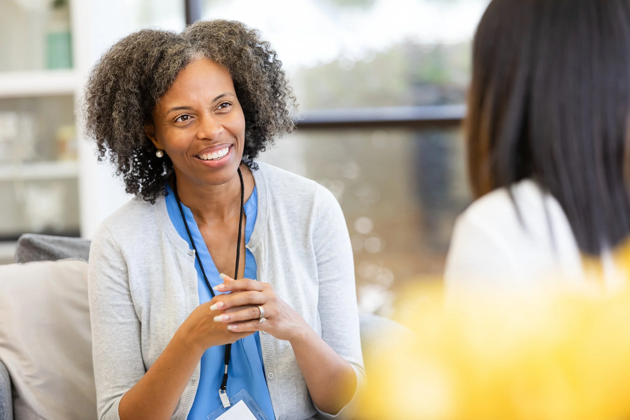 Woman with curly gray hair smiling and talking to another person in a well-lit room.