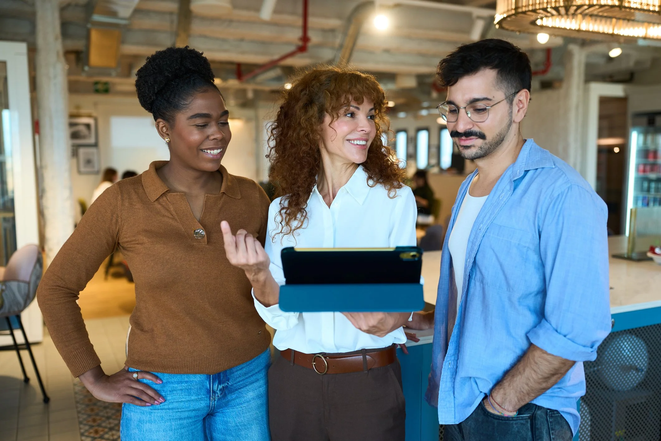 Three people, two women and one man, gathered indoors looking at a tablet device, engaging in a conversation.