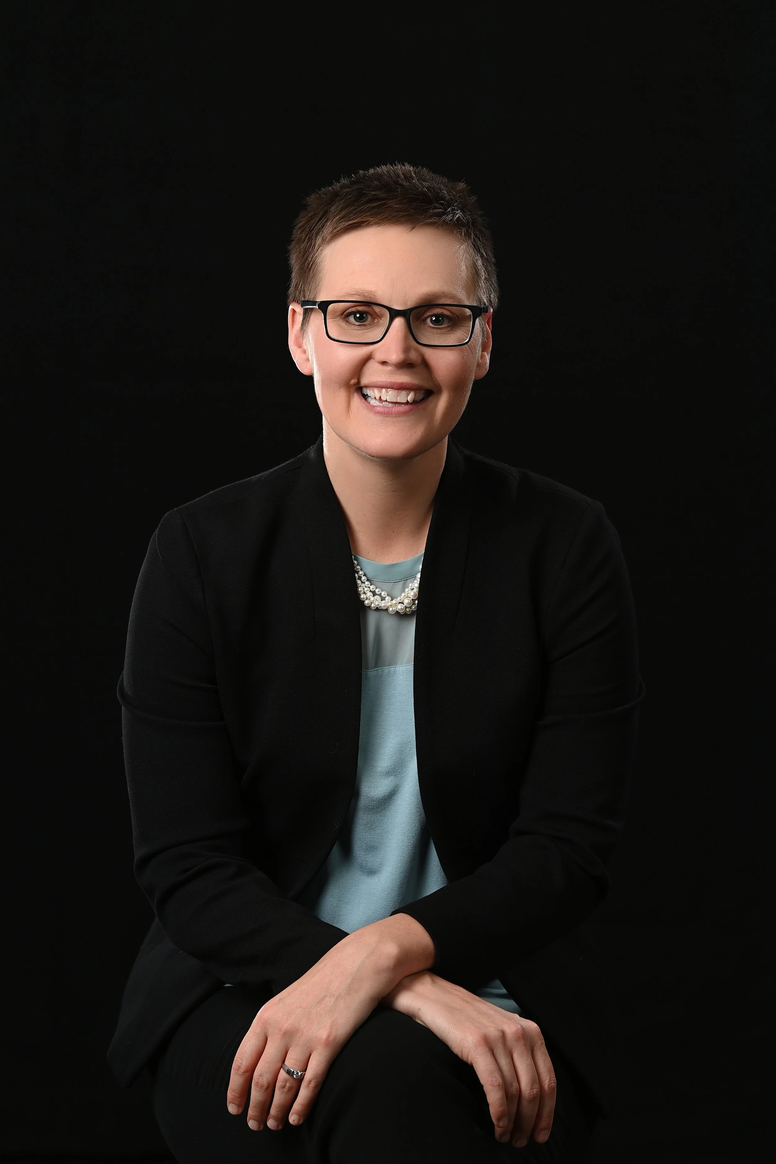 A woman with short hair, wearing glasses, a black blazer, light blue top, and a pearl necklace, sitting against a black background, smiling.