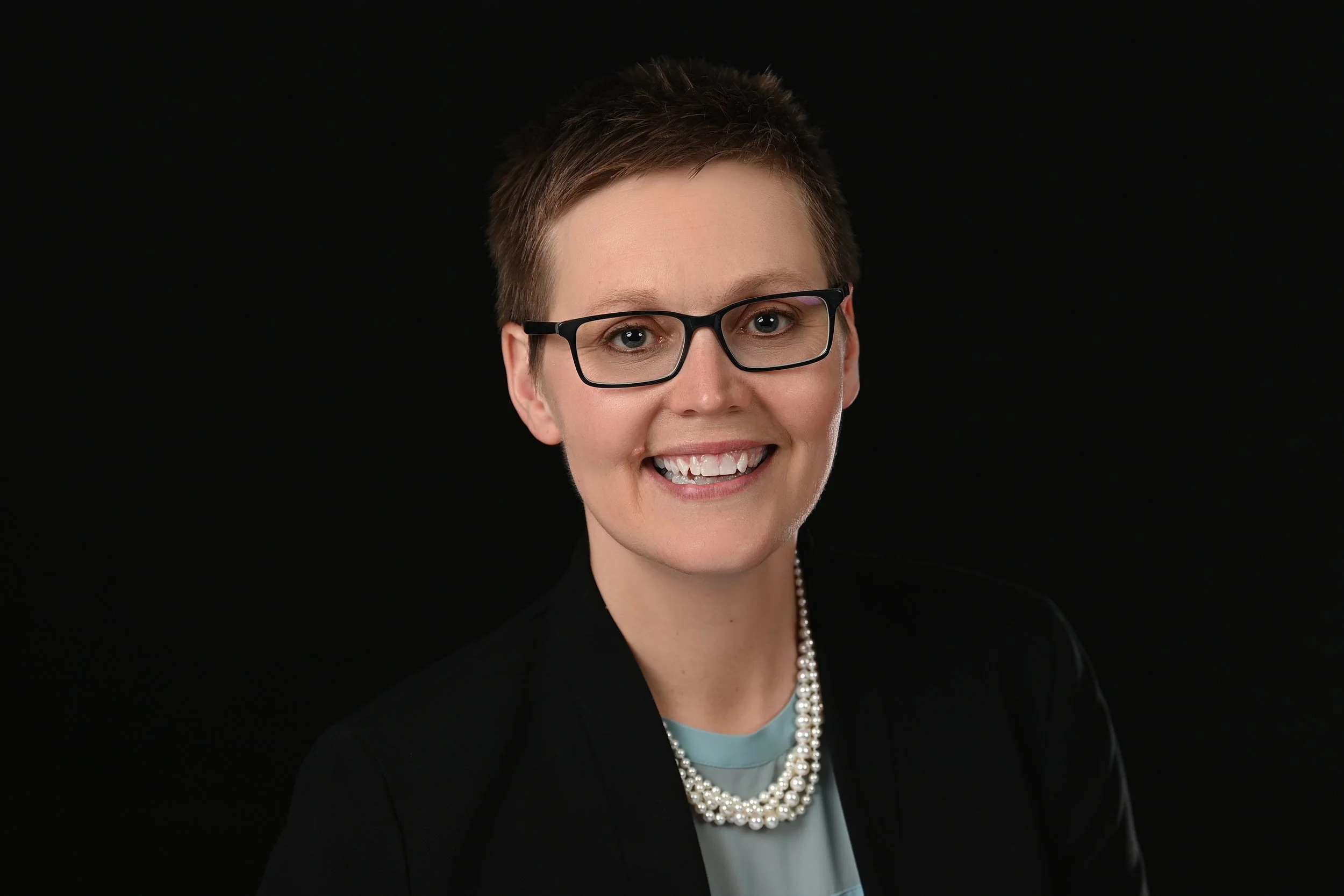 Professional woman with short hair, glasses, wearing a black blazer, pearl necklace, smiling against a black background.
