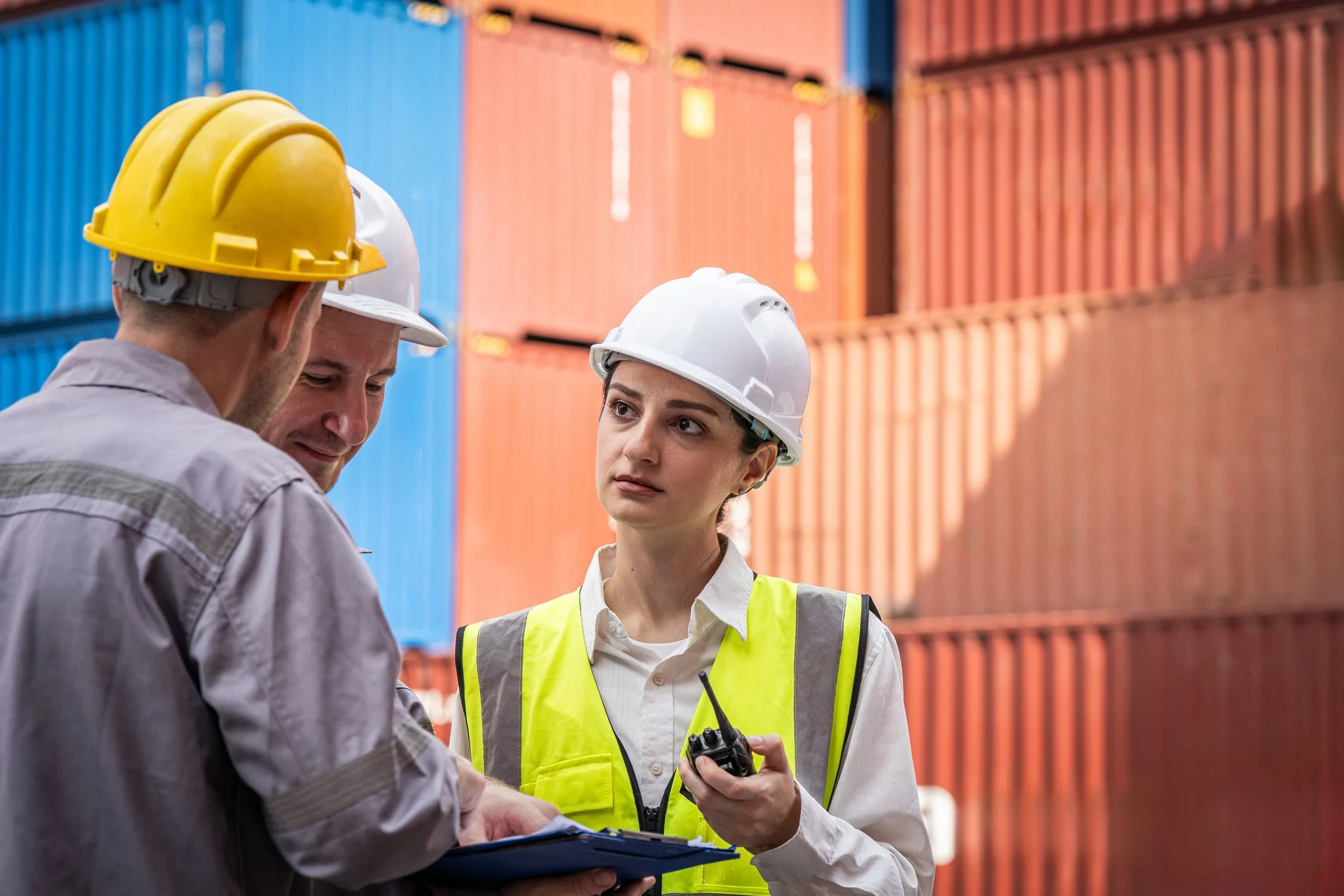Three workers wearing safety helmets and fluorescent vests at a shipping yard with stacked containers.