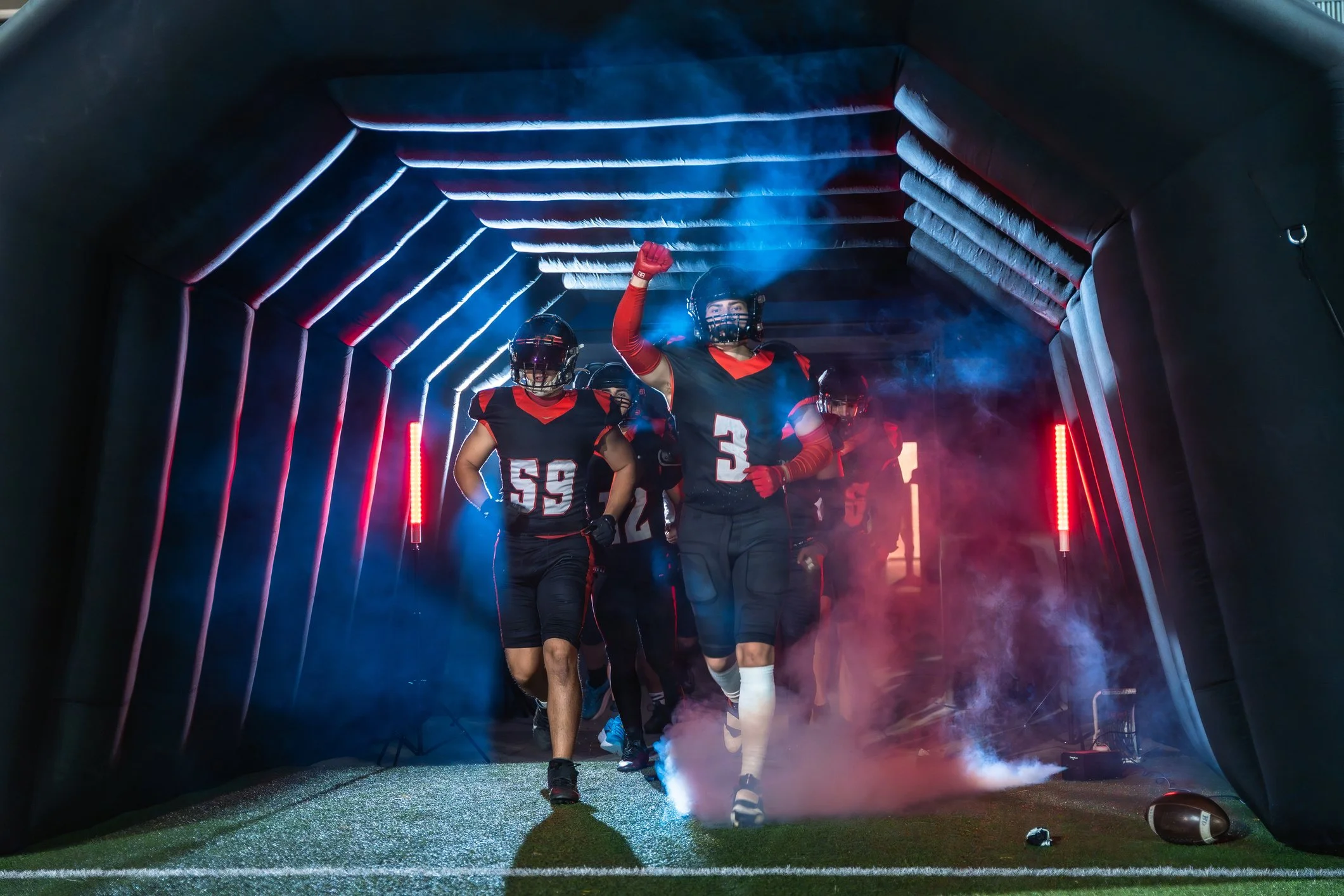 American football players running onto field through inflatable tunnel with smoke and vibrant lighting.